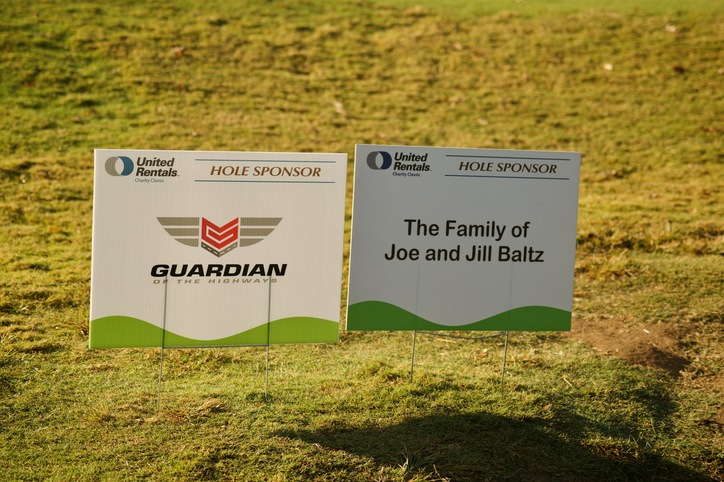 Tournament signage indicating hole sponsors and the family of Joe and Jill Baltz, on a grassy golf course.