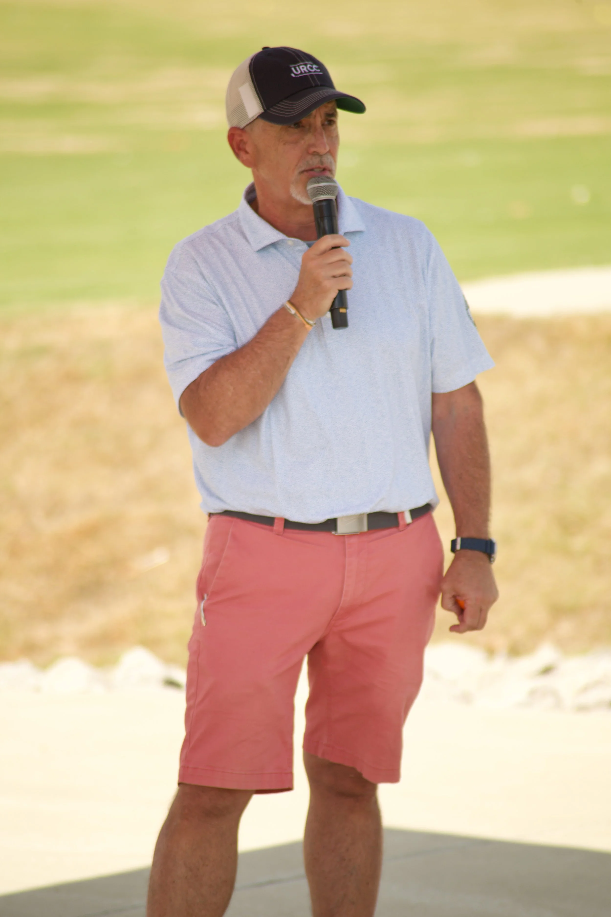 A man wearing a black cap with the letters 'URGE' embroidered on it, a light blue polo shirt, and coral shorts, holding a microphone and standing outdoors on a sunny day.