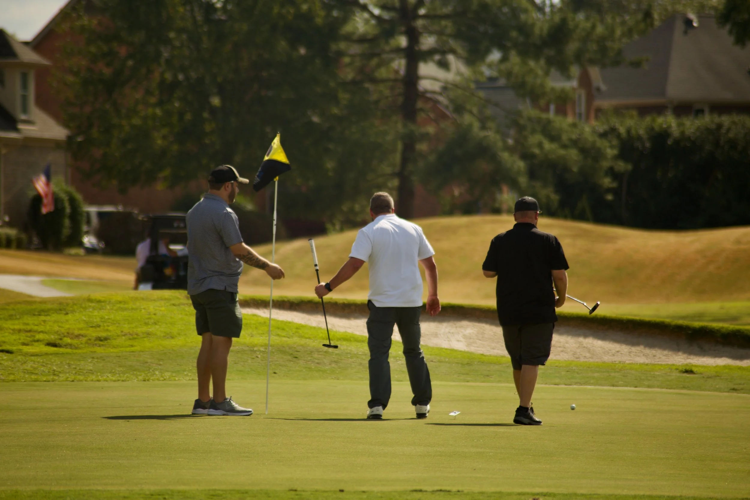Three men on a golf course near a hole, with one holding a golf club, and a golf cart in the background.