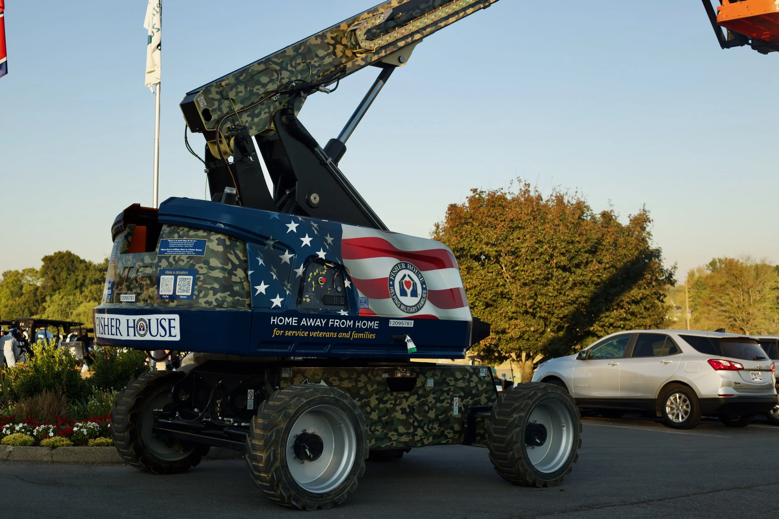 A mobile crane with a camouflage pattern and American flag design, supporting a platform for the Fisher House organization, which assists military veterans and their families, in a parking lot with cars and trees in the background.
