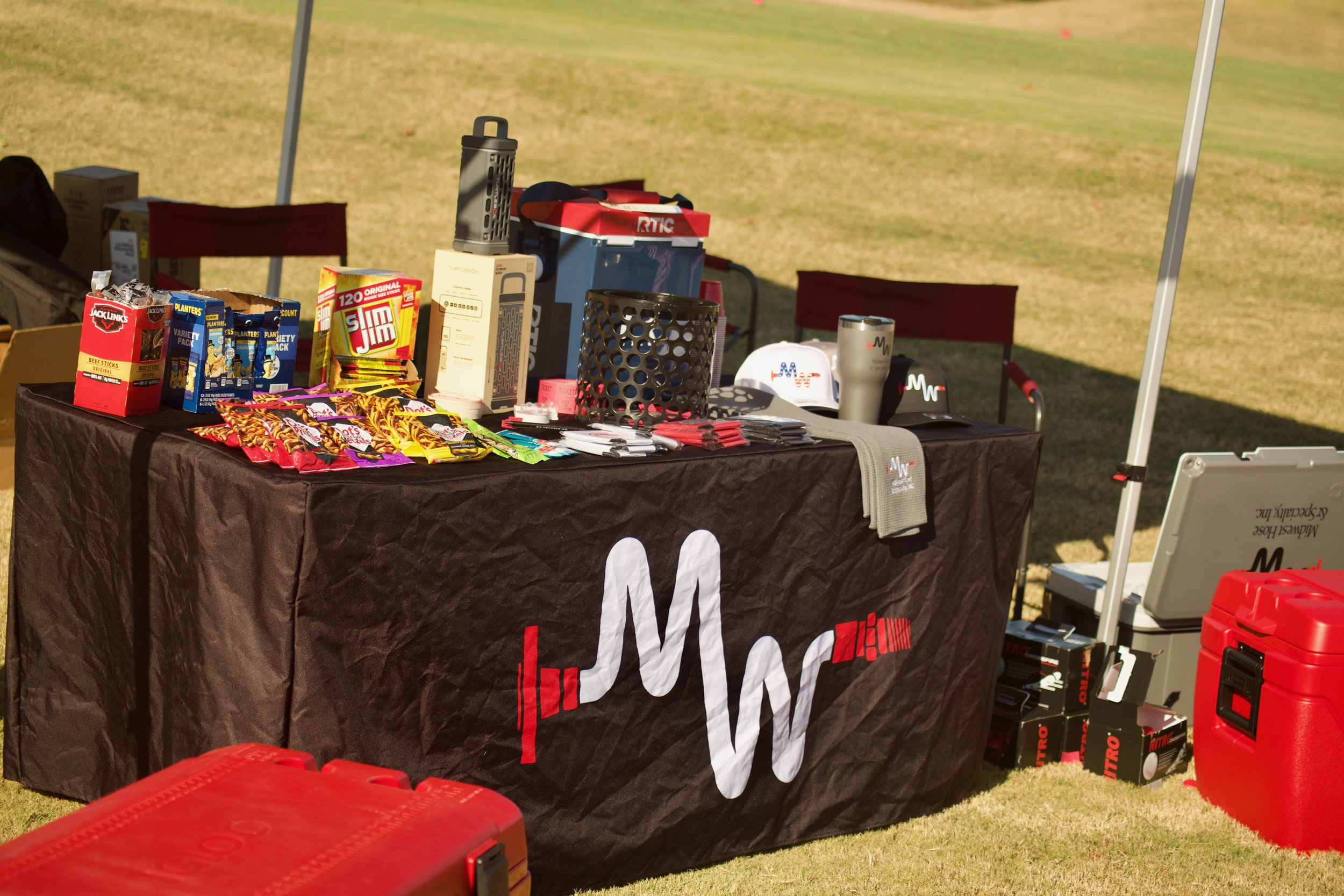 A table covered with a black cloth displaying promotional items including snacks, a water bottle, pens, and a white mug, with a large McDonald's logo and some red boxes in the background, set outdoors on a grassy area.