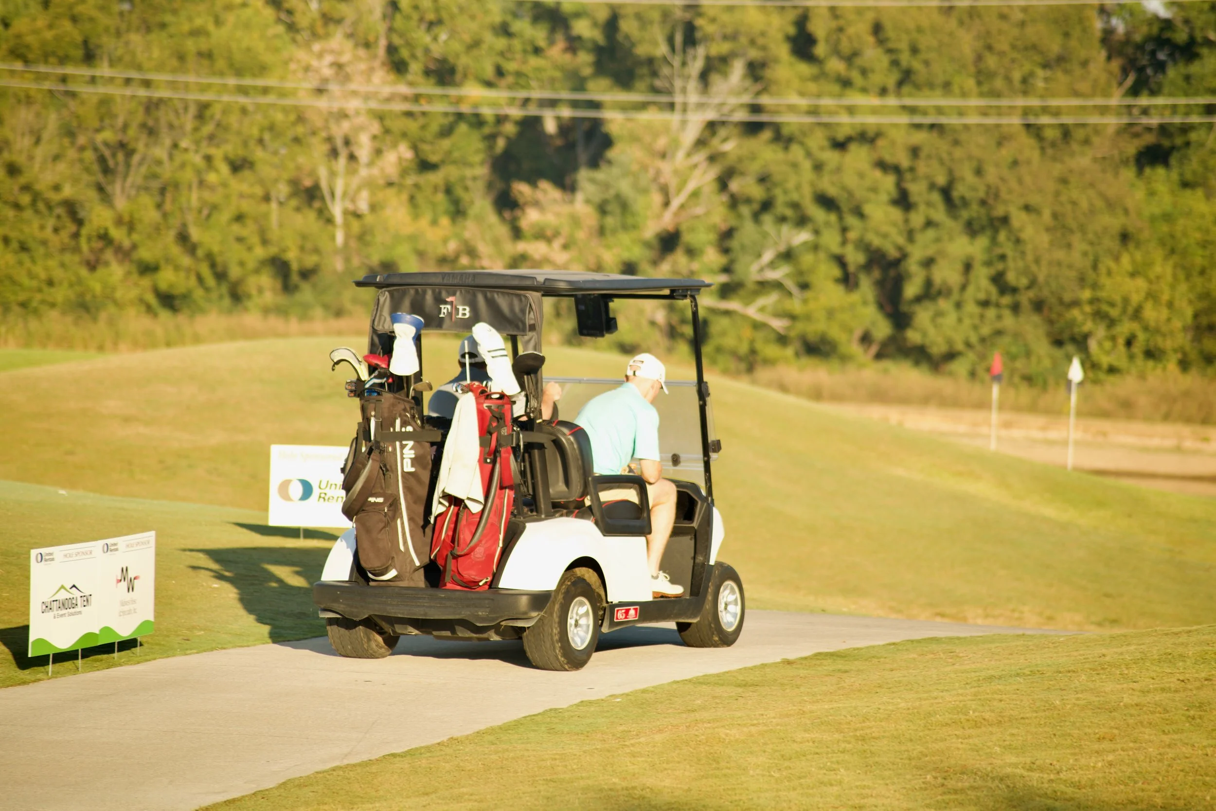 A golf cart with two men inside, one driving and the other seated, on a golf course with trees in the background and flags on the greens.