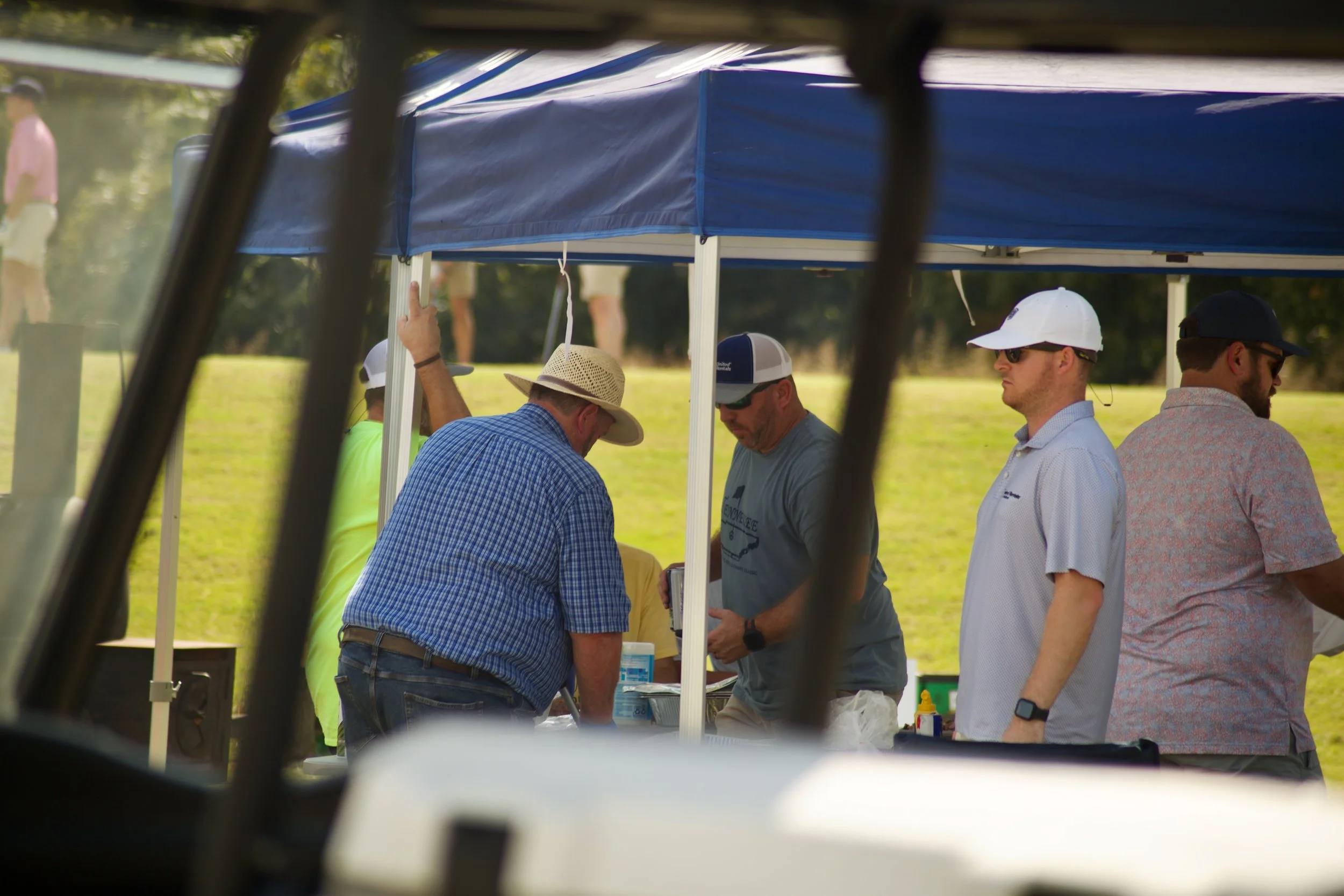People gathered under a blue canopy tent at an outdoor event, viewed from inside or through an opening of a vehicle or structure, with some individuals wearing caps and sunglasses, and engaging in conversation.