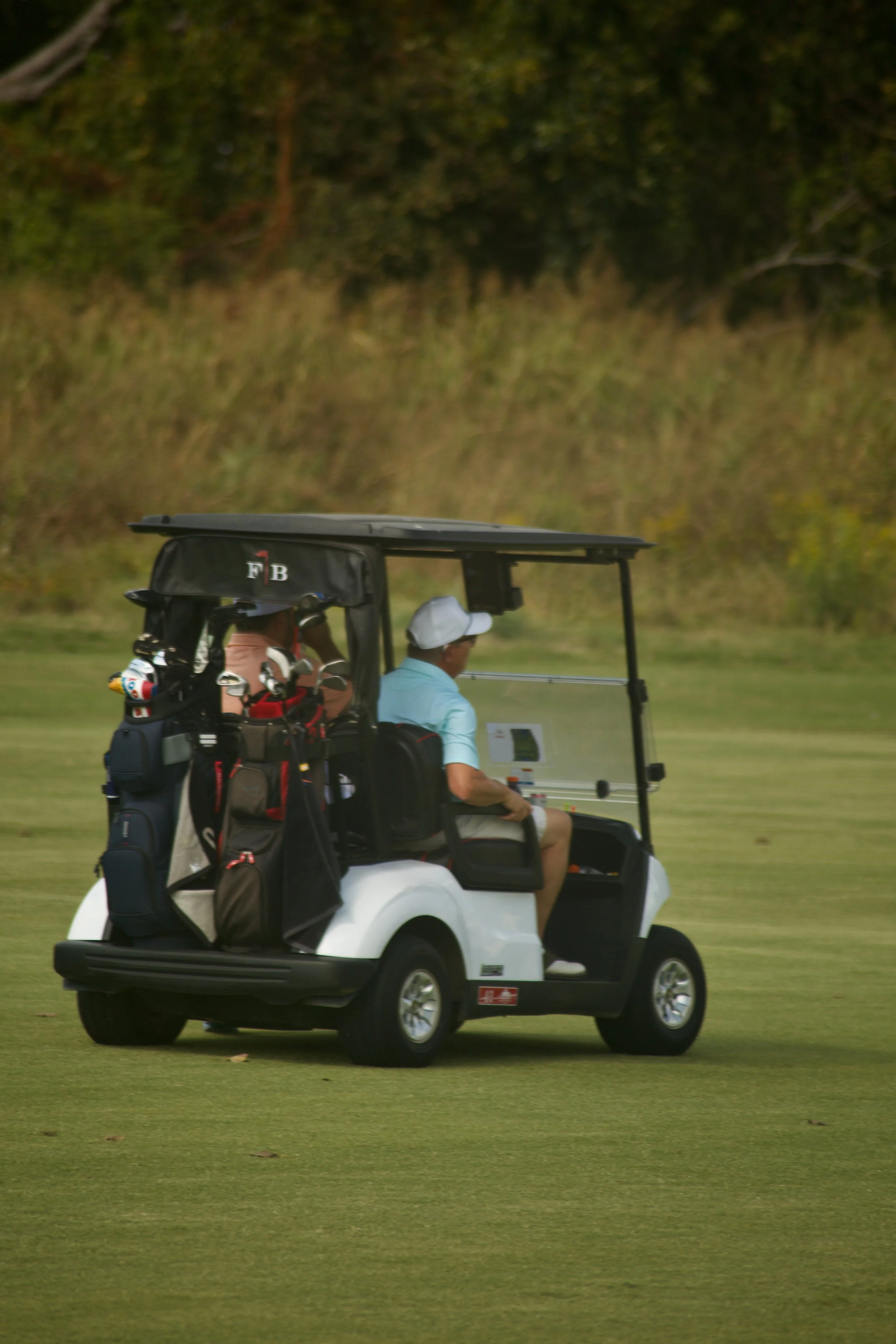 A man and a woman riding in a golf cart on a green golf course, with trees in the background.