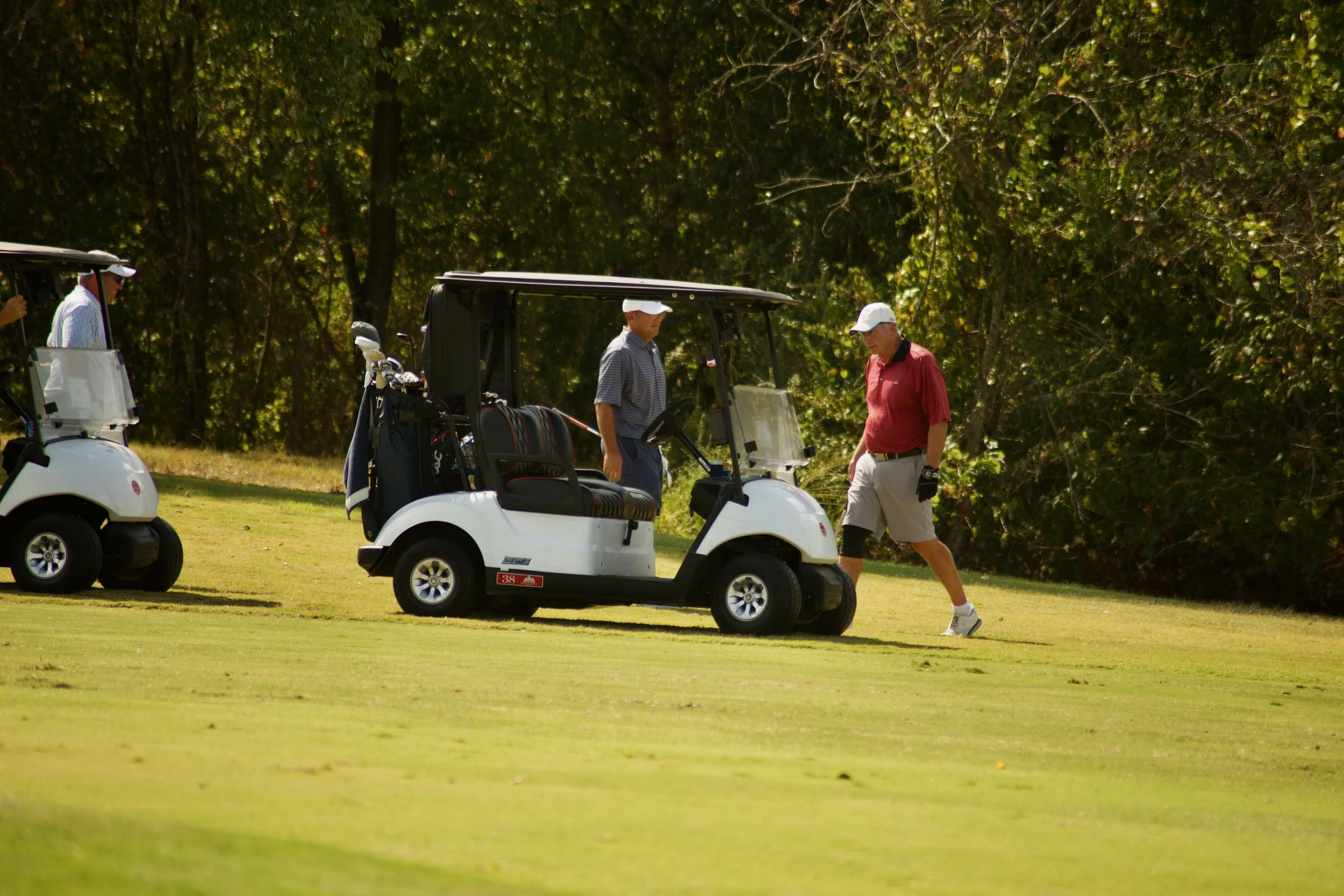 Two men playing golf, one in a red shirt and white cap, the other in a striped shirt and white cap, near a golf cart on a green golf course with trees in the background.