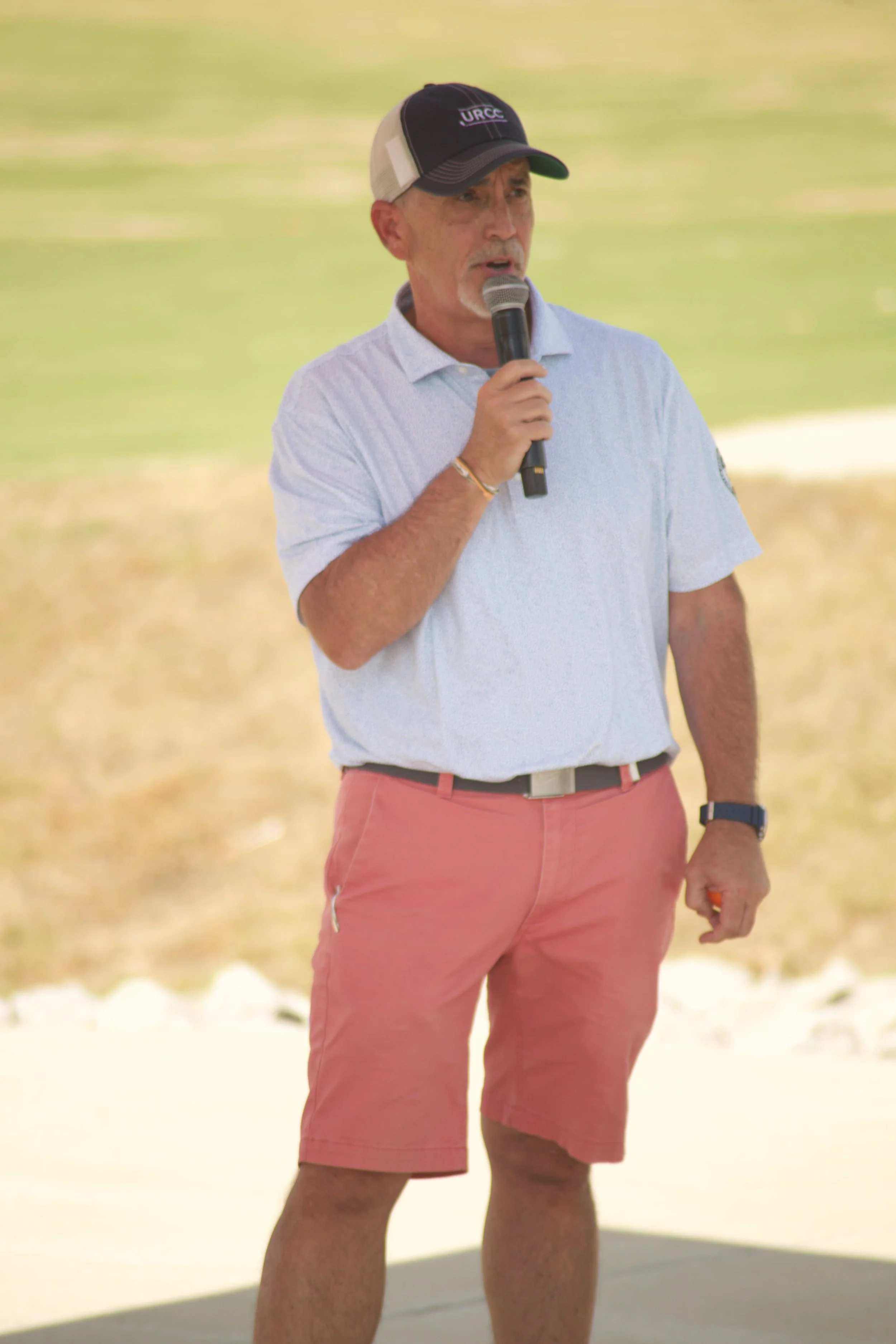 A man standing outdoors near a body of water, speaking into a microphone, wearing a light blue shirt, pink shorts, a black and white cap, and a watch on his wrist.