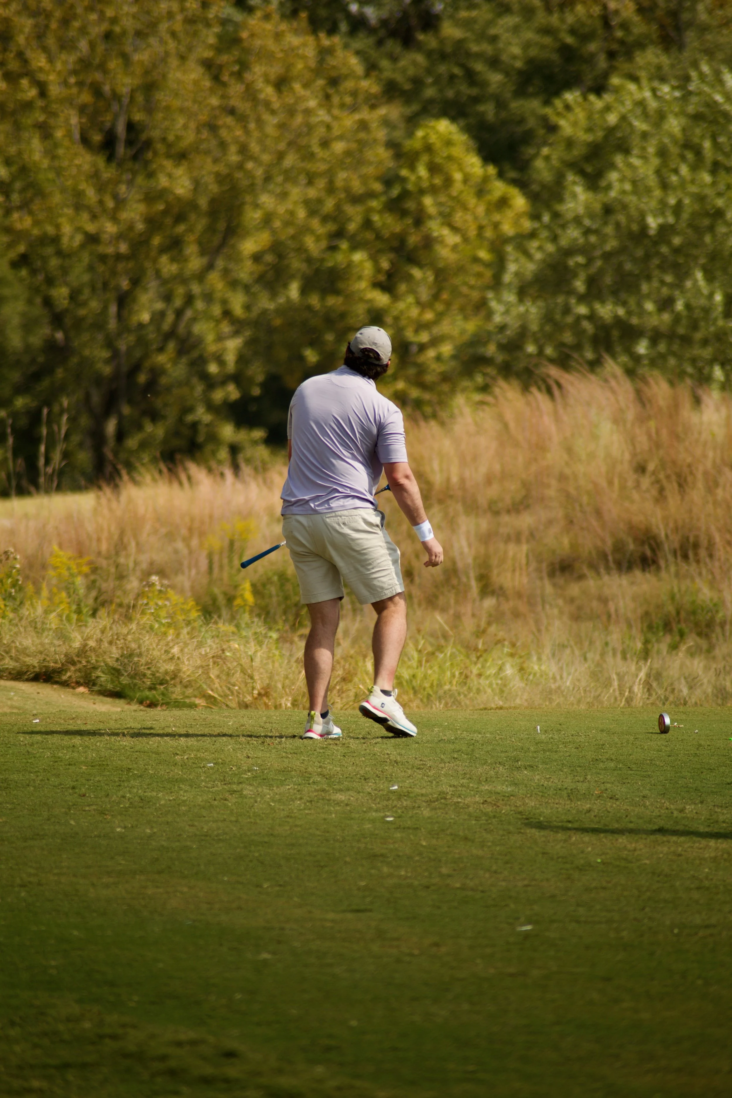 A man on a golf course preparing to hit a golf ball, wearing a gray cap, purple shirt, beige shorts, and white golf shoes, with trees and grass in the background.