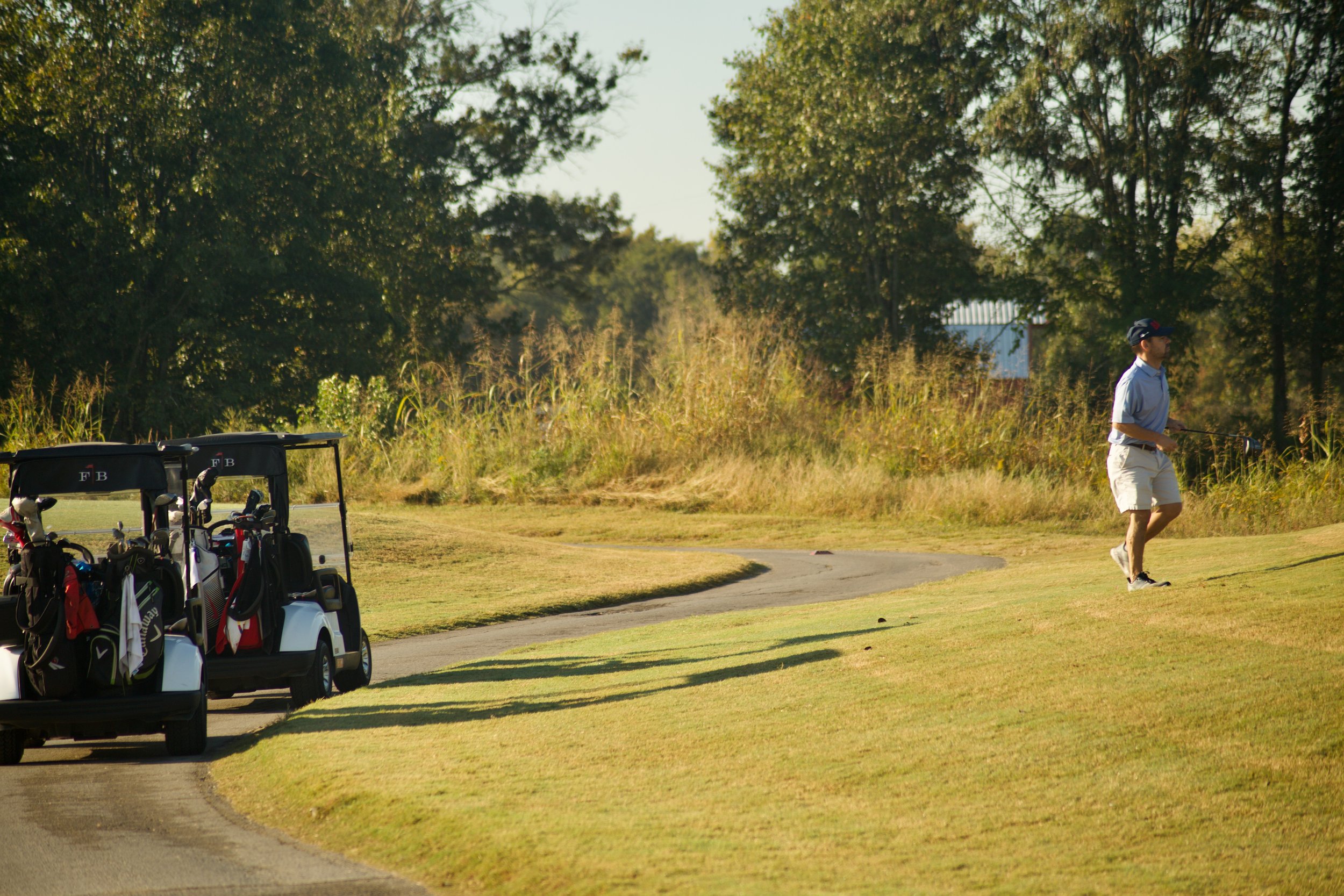 A person in a light blue shirt and beige shorts walking on a golf course with a golf club in hand, with golf carts and bags visible in the background, surrounded by trees and greenery.