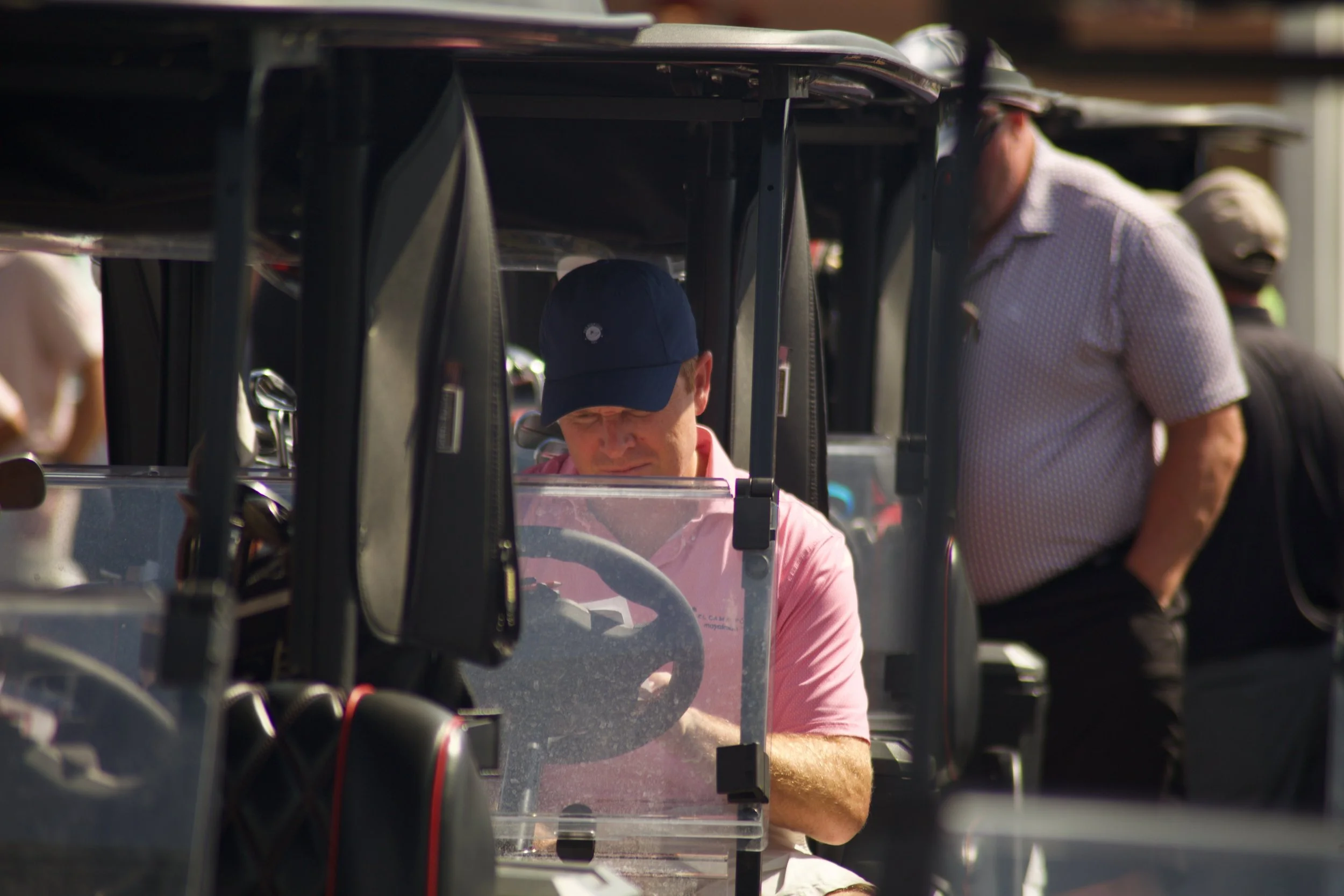 A person with a pink shirt and a blue baseball cap is sitting in a golf cart, with another person leaning over talking to them, in an outdoor setting with multiple golf carts around.
