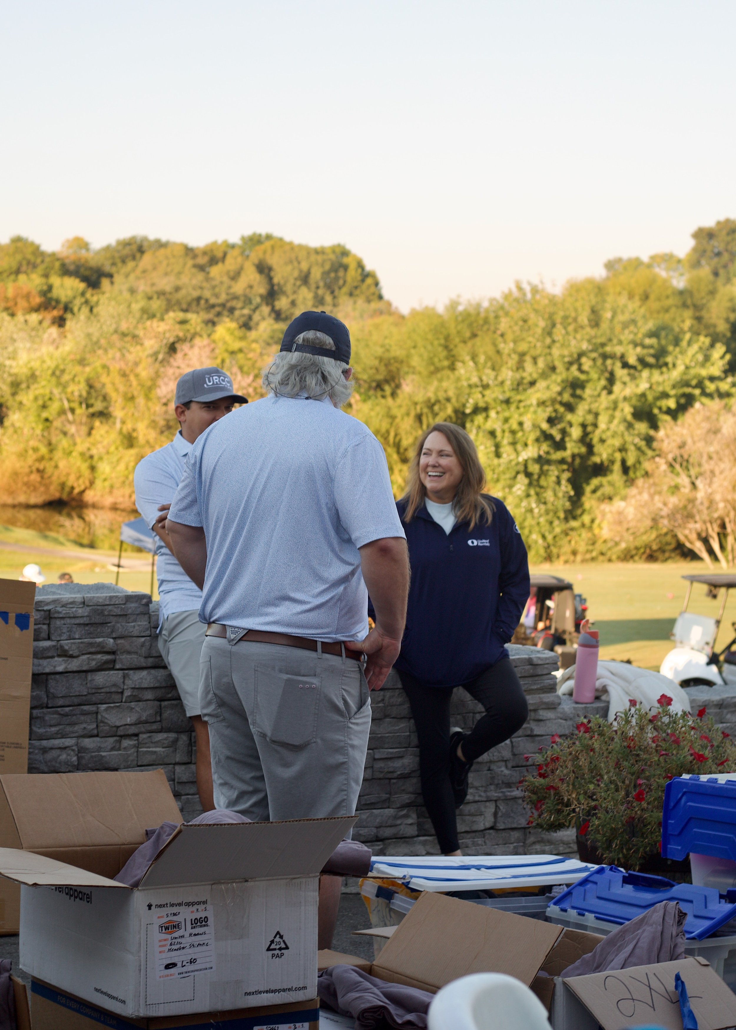 People talking at an outdoor event near a river, with trees in the background. There are boxes and supplies in the foreground.