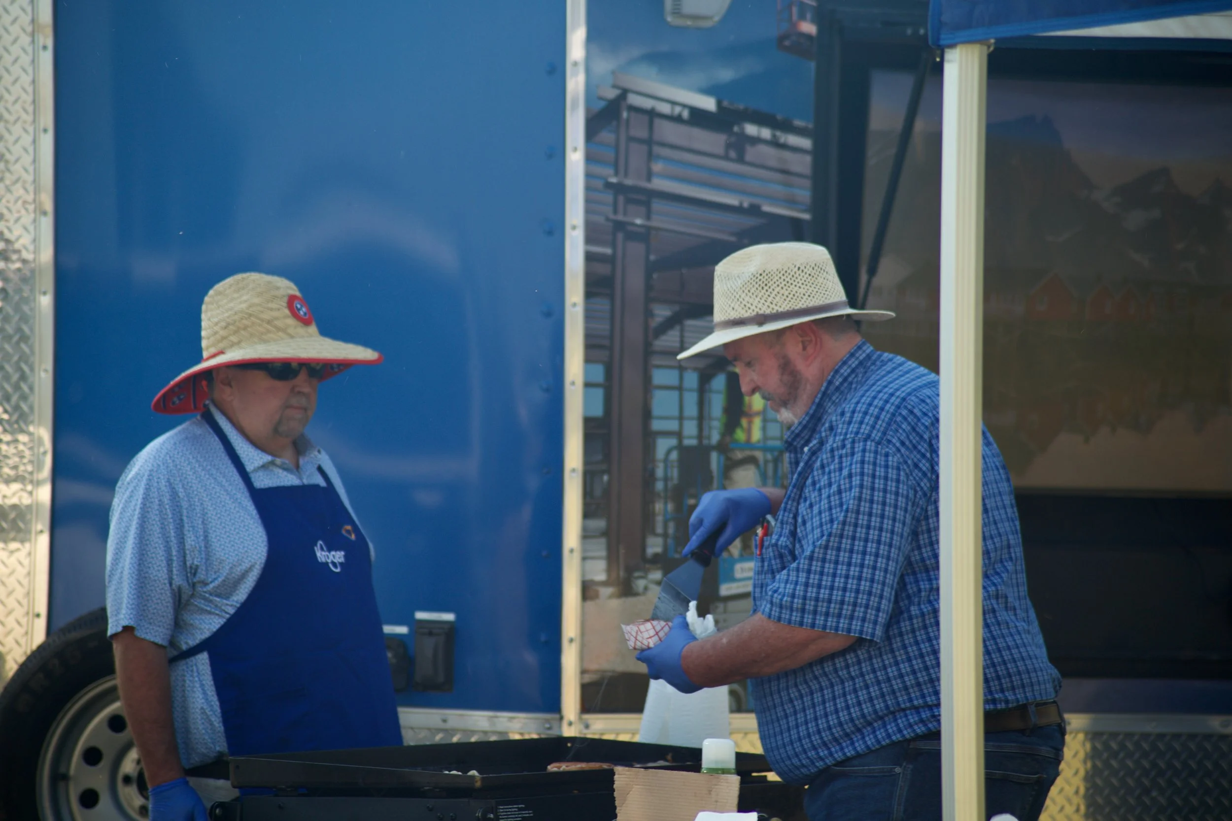 Two men wearing hats and aprons standing at a food truck, with one man preparing food and the other observing. They are outdoors on a sunny day.