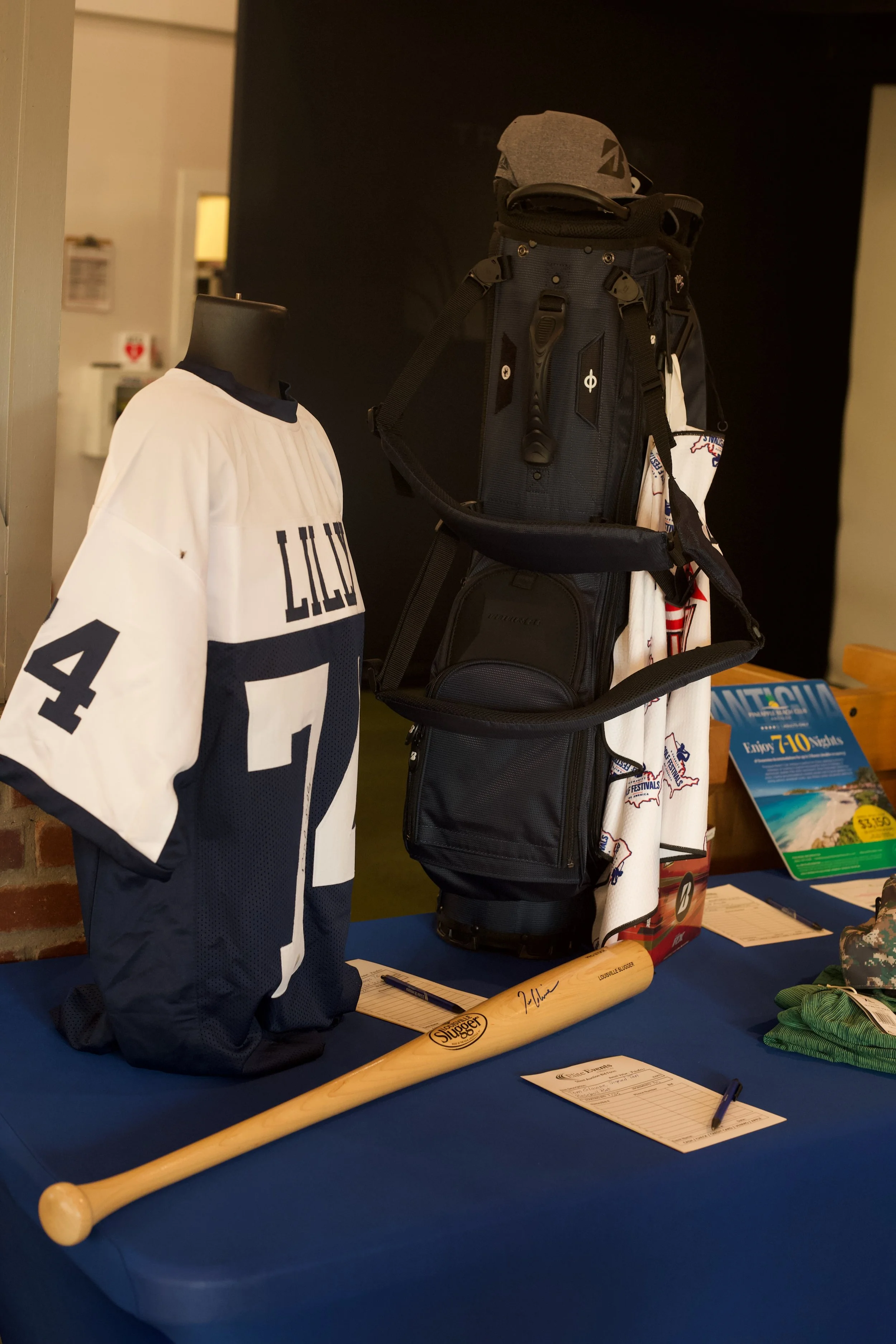 Display of sports memorabilia, including a white and navy jersey with the number 4, a signed baseball bat, a black backpack with hockey equipment, and a sign about travel brochures on a table covered with a blue cloth.