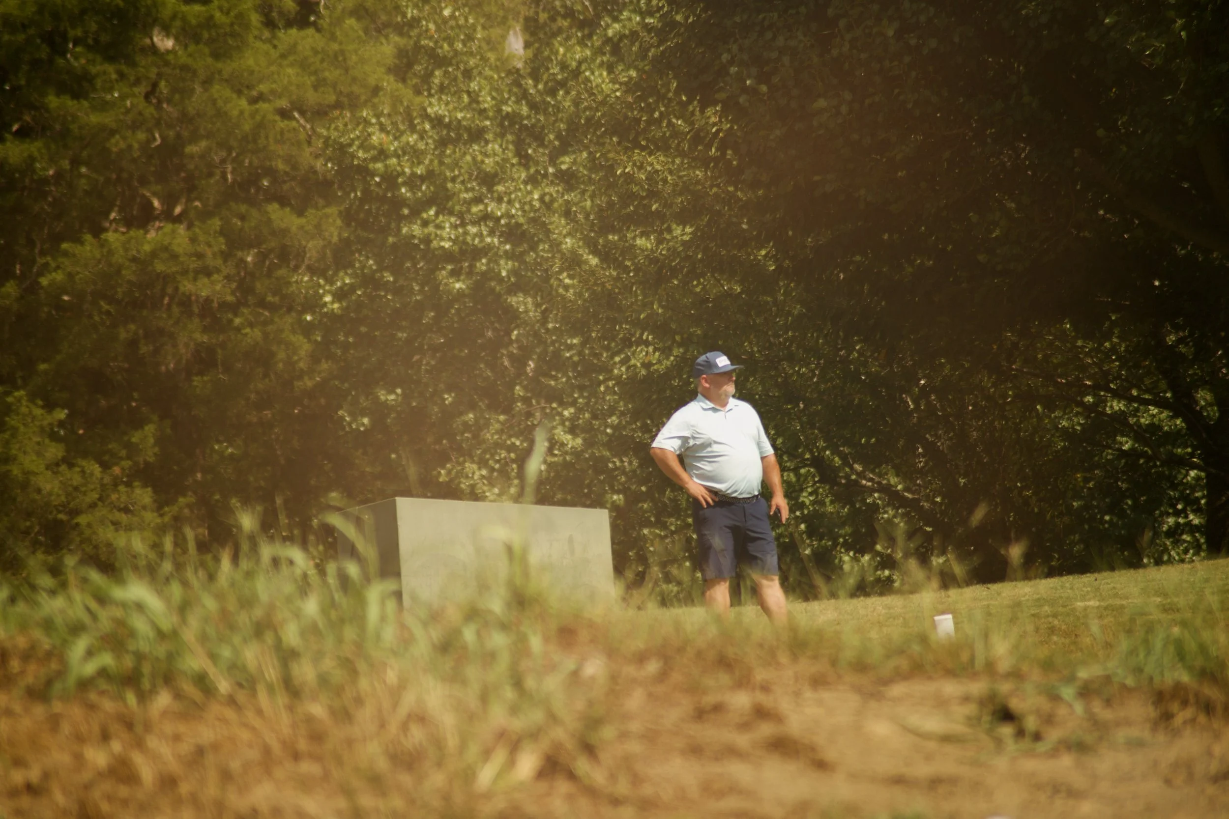 A man wearing a light-colored shirt, dark shorts, and a cap standing outdoors near greenery and a concrete structure.