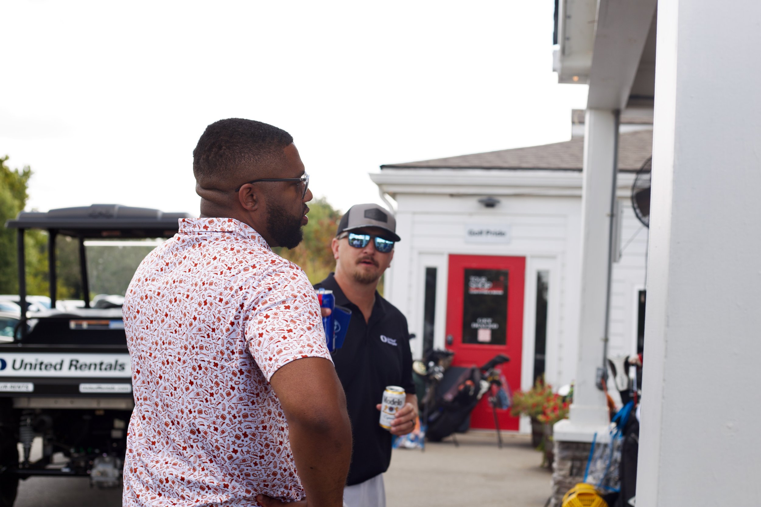 A man with glasses and a beard wearing a white shirt with red patterns, holding a can, standing outdoors near a food truck and a red fire alarm box, with another person in the background wearing sunglasses and a hat.