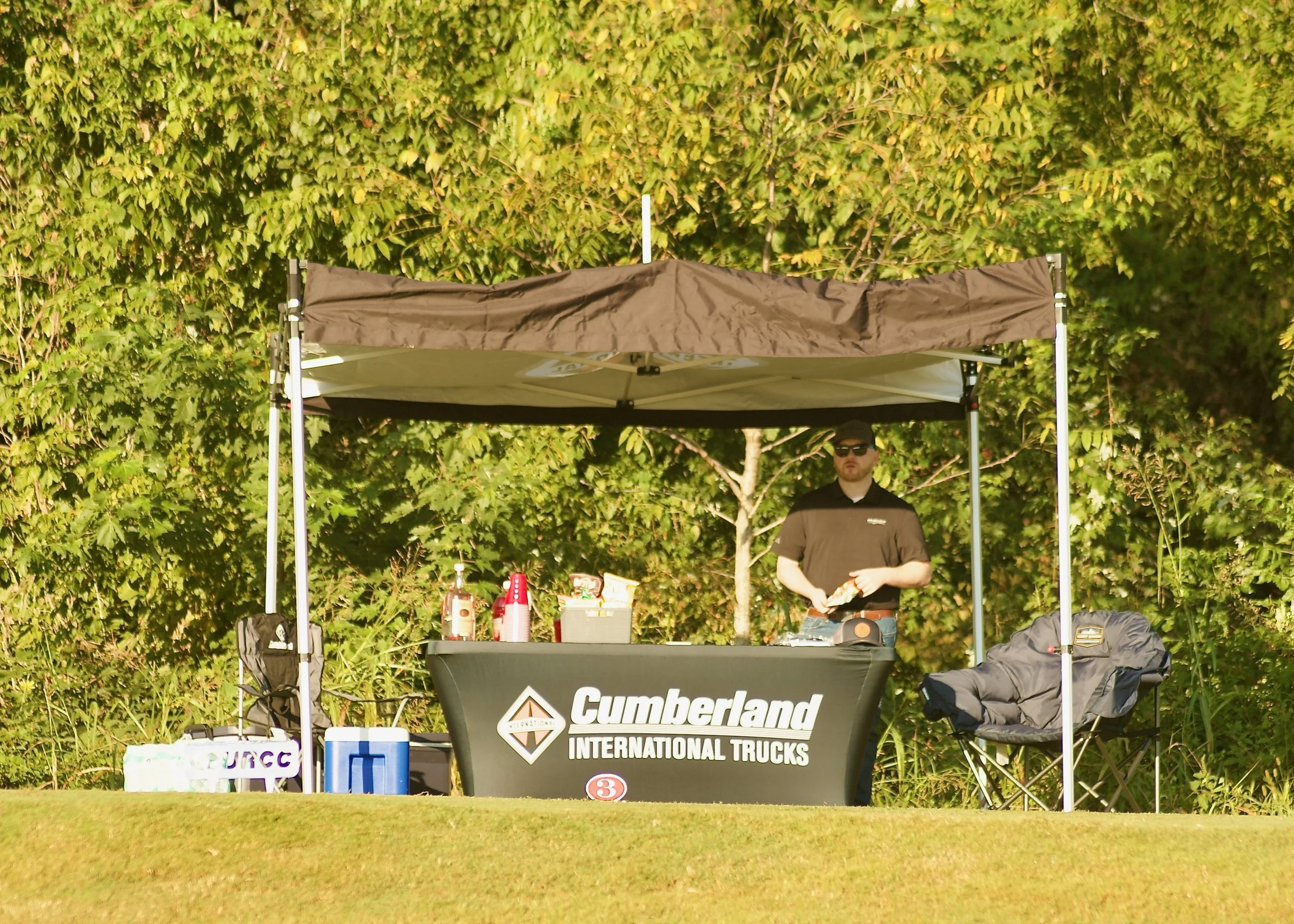 A man wearing sunglasses and a black Mountain Dew jacket standing behind a black Cumberland International Trucks table at an outdoor event with a green leafy hedge in the background.