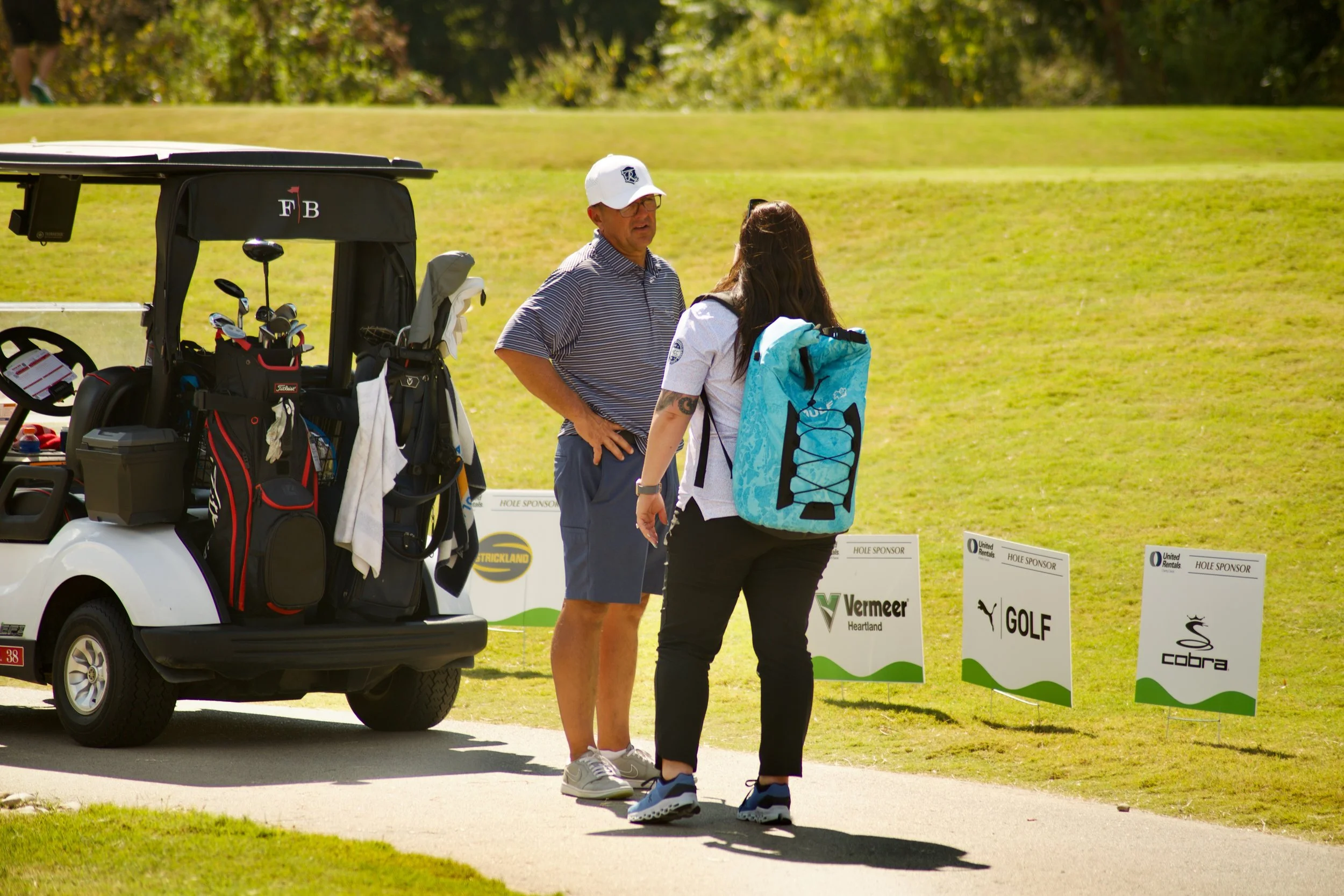 A man and a woman stand on a golf course near a driving range cart filled with golf clubs. The man wears a white cap, striped polo, and glasses, while the woman has long dark hair, a white shirt, black pants, and a blue golf bag. They are talking, wi