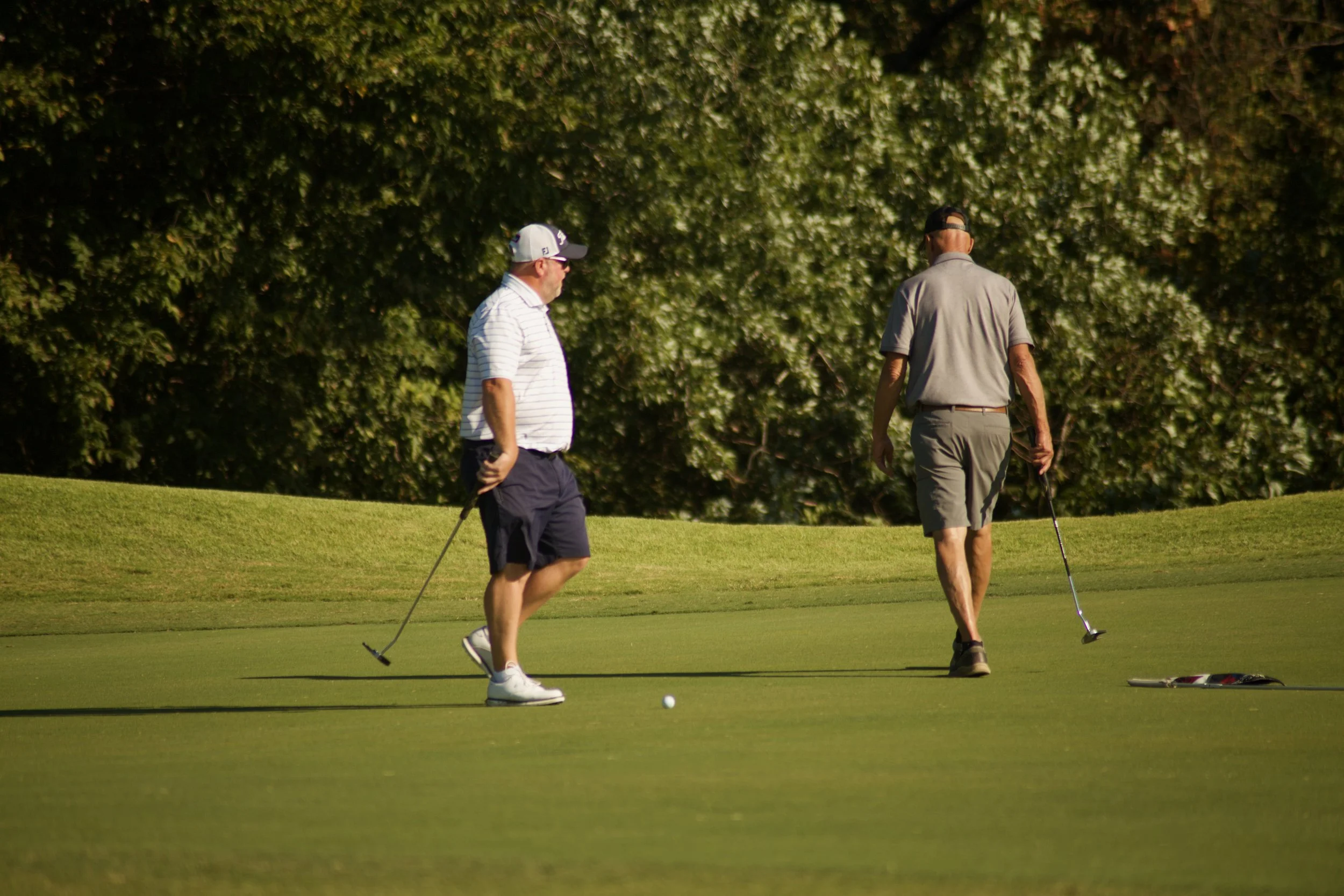 Two men playing golf on a course, with one preparing to putt and the other watching, set against a backdrop of lush green trees.