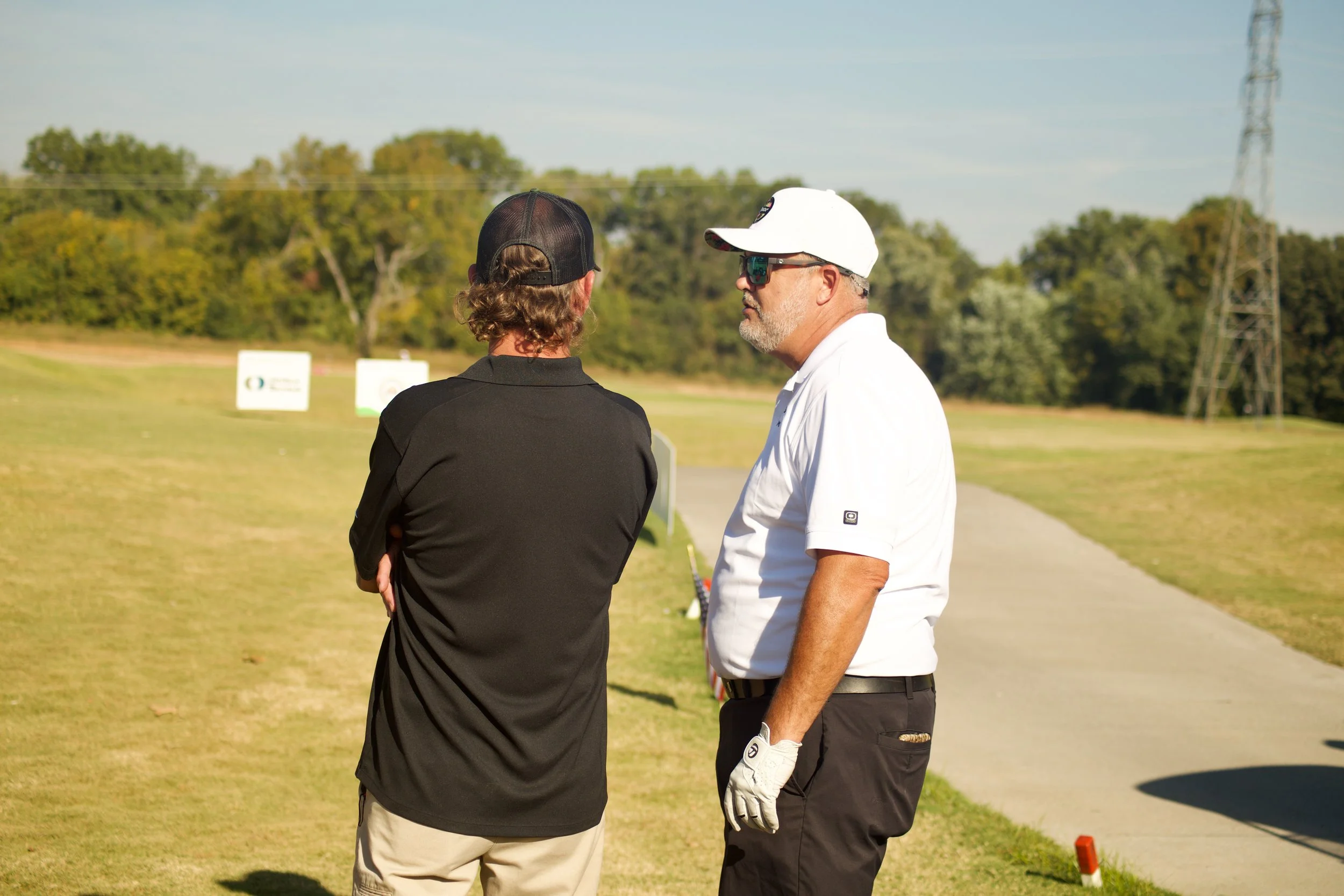 Two men standing and talking outdoors at a golf course, one wearing a black shirt and cap, the other in a white polo and cap, with trees and signs in the background.