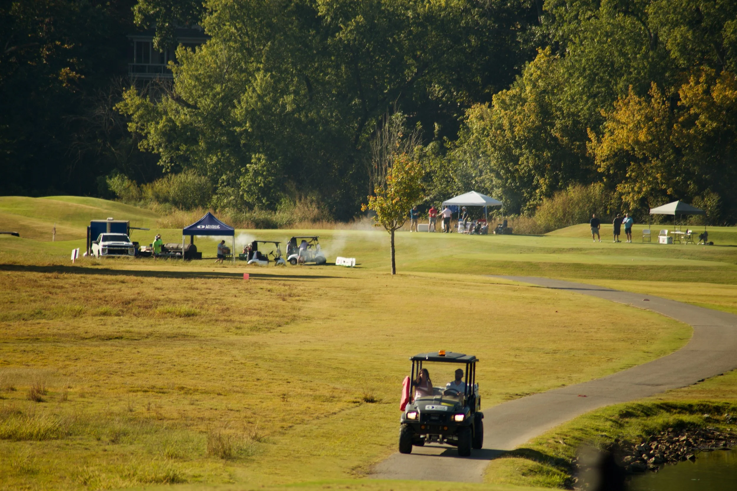 A golf course with people playing, tents set up, and golf carts on the grass and a paved path, surrounded by tall green trees.