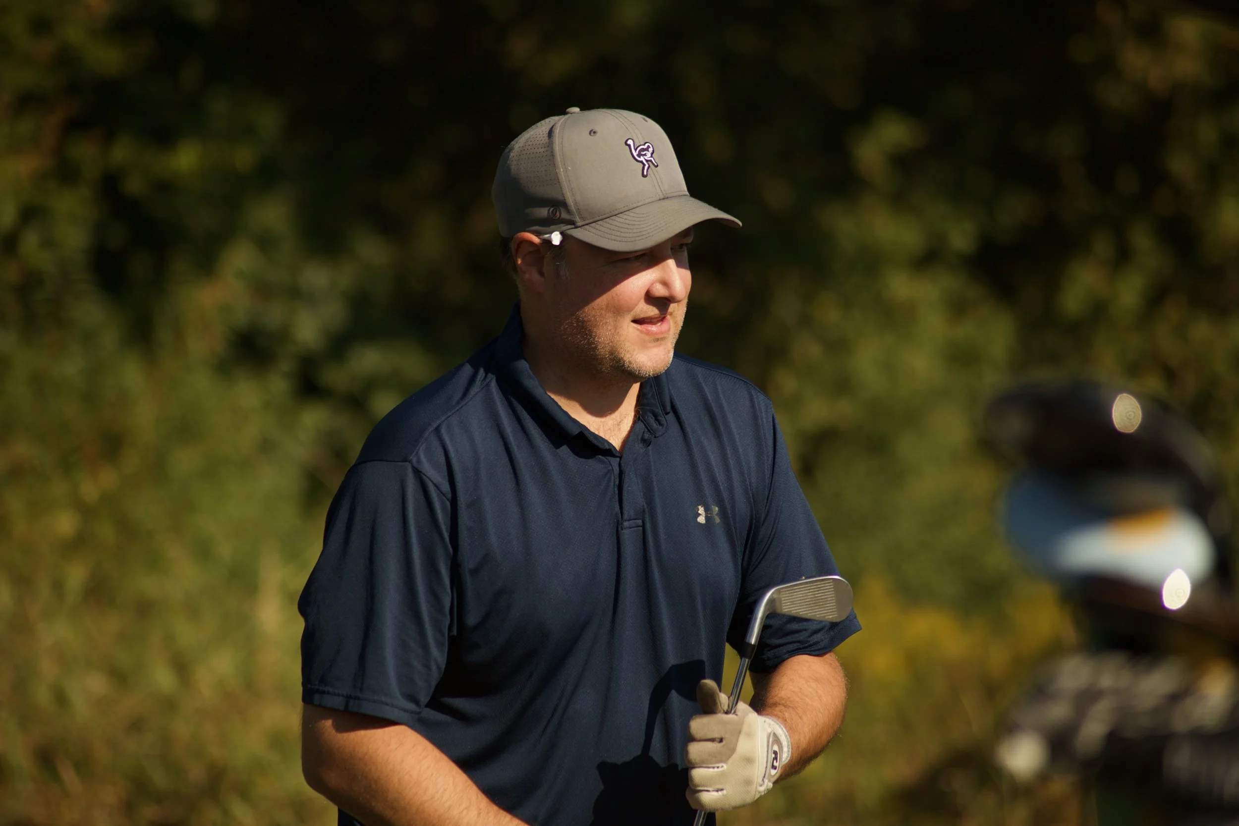 A man wearing a light gray baseball cap with a purple flamingo logo, a dark navy Under Armour shirt, and sunglasses, holding a golf club and standing outdoors with blurry green trees in the background.