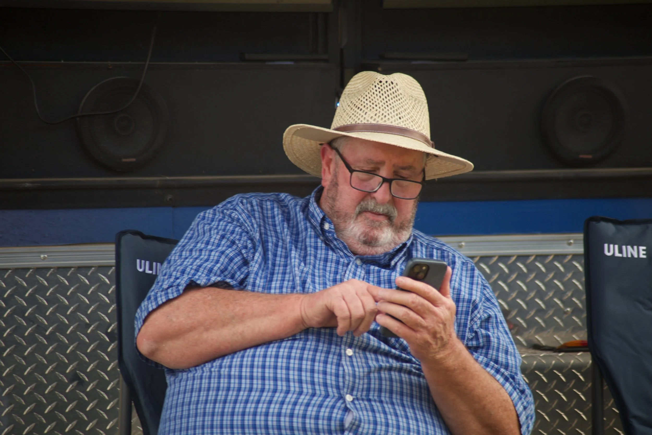An older man with glasses and a beard, wearing a straw hat and a blue checkered shirt, is sitting outdoors and looking at his smartphone. He is seated in a folding chair with ULINE branding, with a metal diamond plate surface behind him and two black