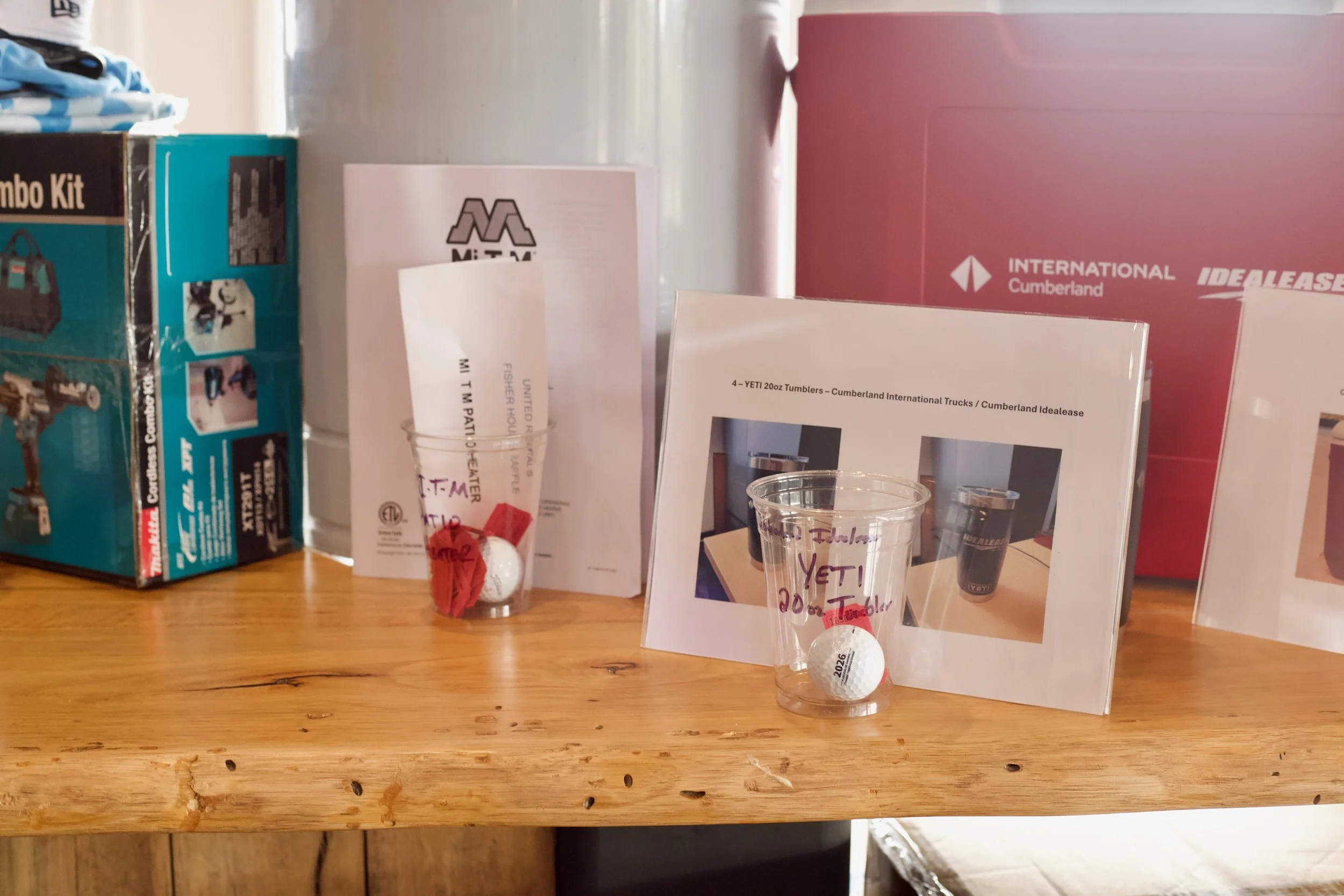 Display of various items on a wooden table, including boxes, pamphlets, and plastic cups with golf balls inside, labeled for a tournament event.