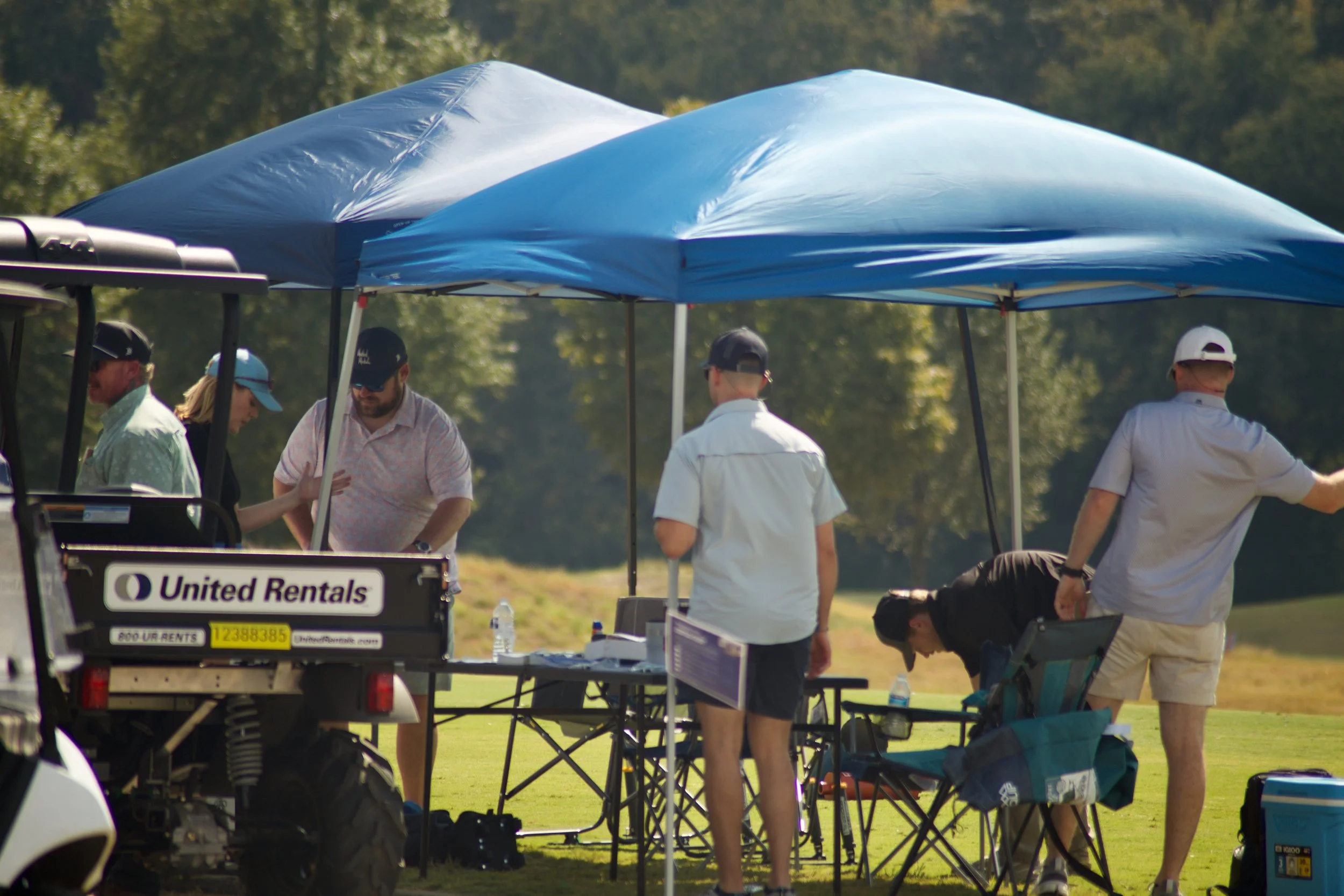 People gathered under a blue canopy tent at an outdoor event with equipment and supplies, including a golf cart labeled 'United Rentals' in the foreground.