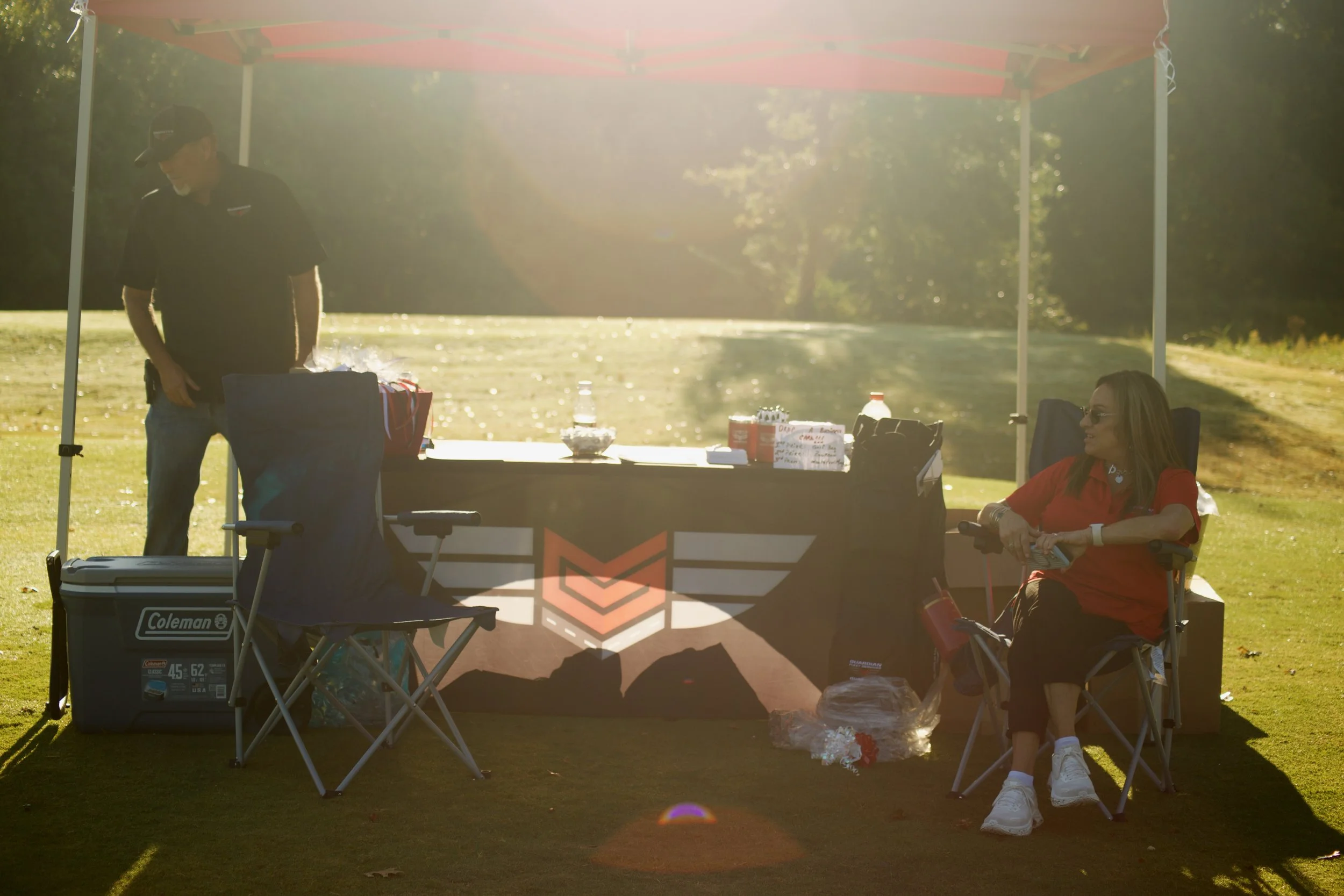 An outdoor scene with a man standing and a woman sitting under a canopy on chairs. The man is near a table with snacks and drinks, and the woman is wearing sunglasses and a red shirt. Sunlight is shining brightly, creating a lens flare effect.