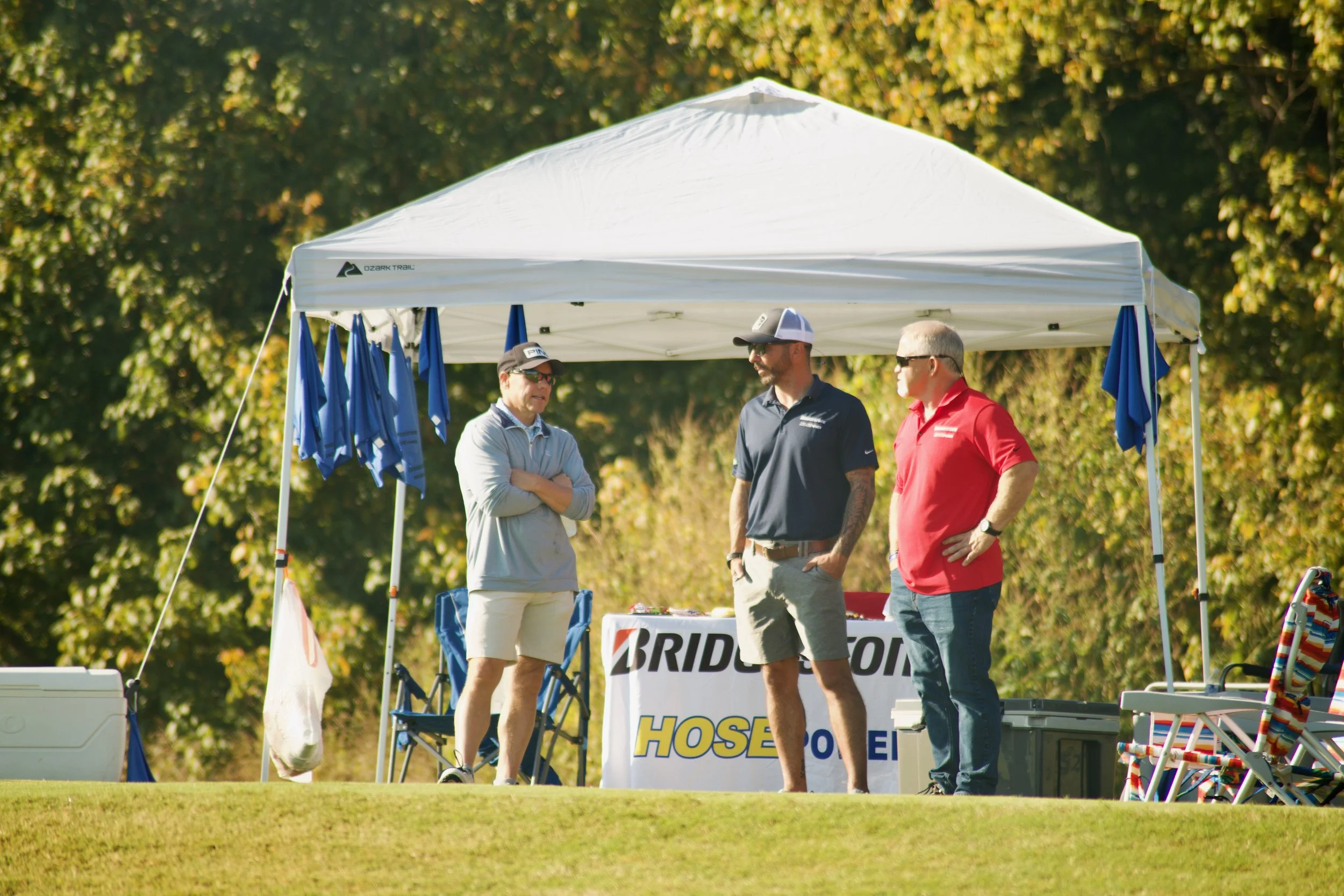 Three men stand under a white canopy tent outdoors on a sunny day, engaging in conversation. The tent has blue umbrellas hanging on it, and there are chairs and tables nearby. The background features trees with green and yellow leaves.