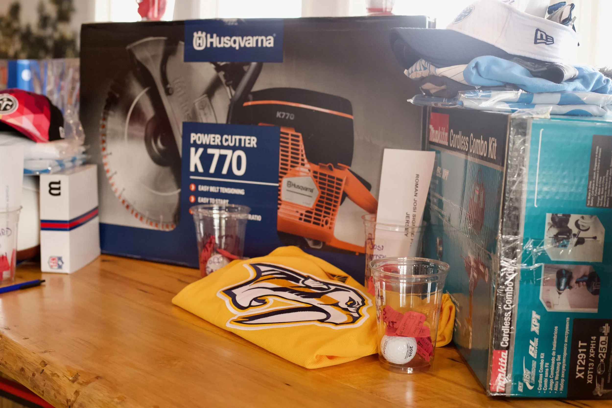 Various sports and outdoor equipment including a Husqvarna power cutter, a TaylorMade golf ball, a Denver Broncos hat, and other boxed items on a wooden table.