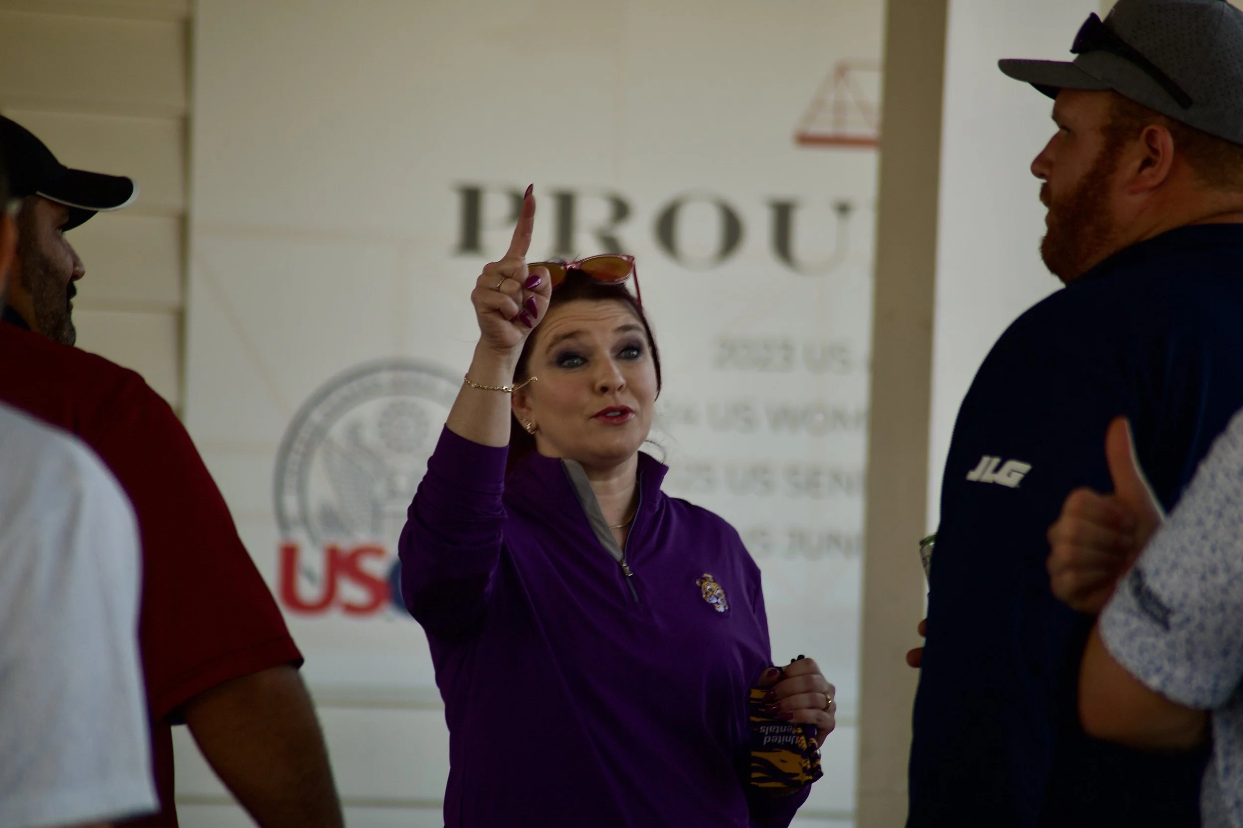 A woman with red hair and purple jacket holds up her index finger and appears to be speaking to two men, one on each side, in a setting with a sign that reads 'PROU' in the background.