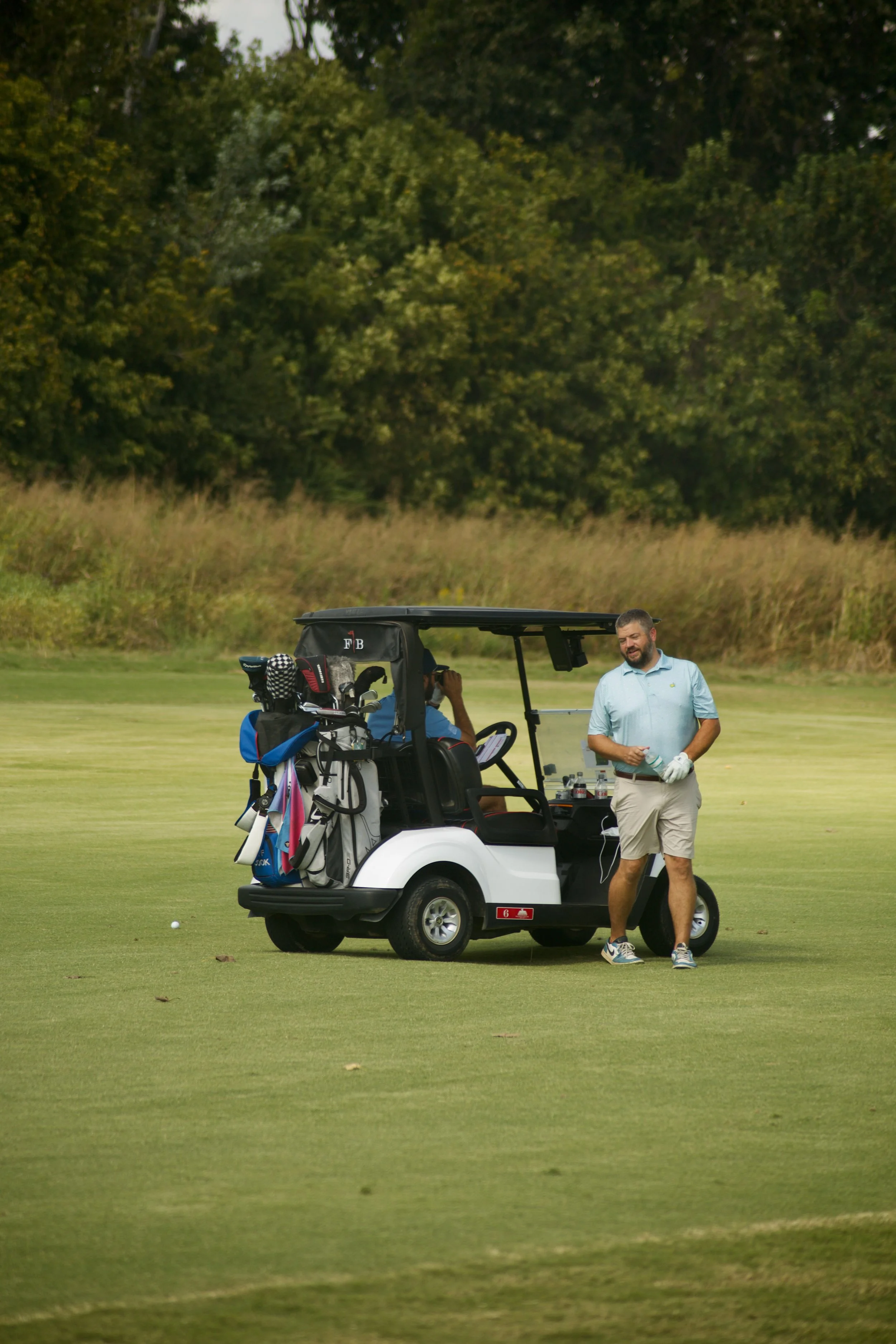 A man standing near a golf cart on a golf course, holding a golf glove and a water bottle, with a golf bag and clubs in the cart.