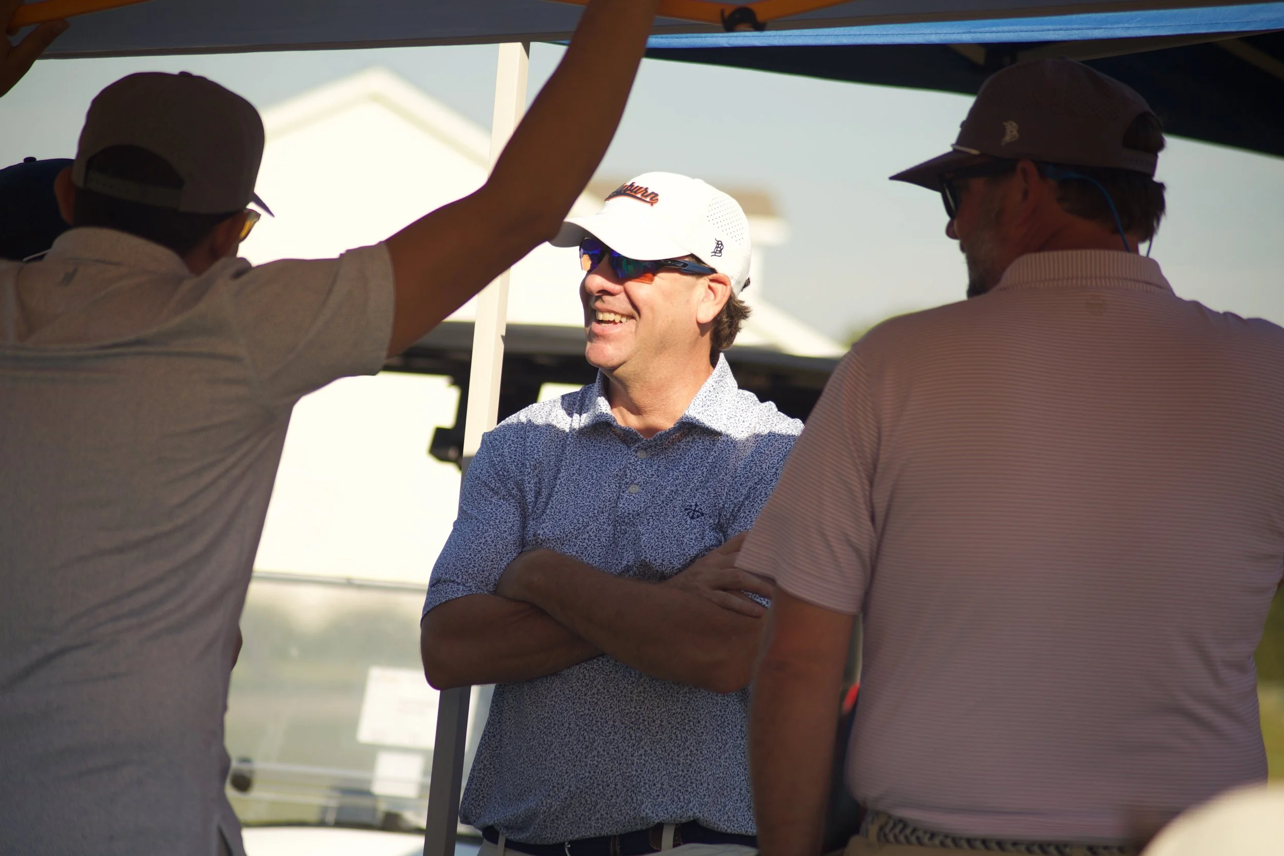 Three men in casual clothing and hats standing under a canopy, smiling and engaging in conversation during daytime.