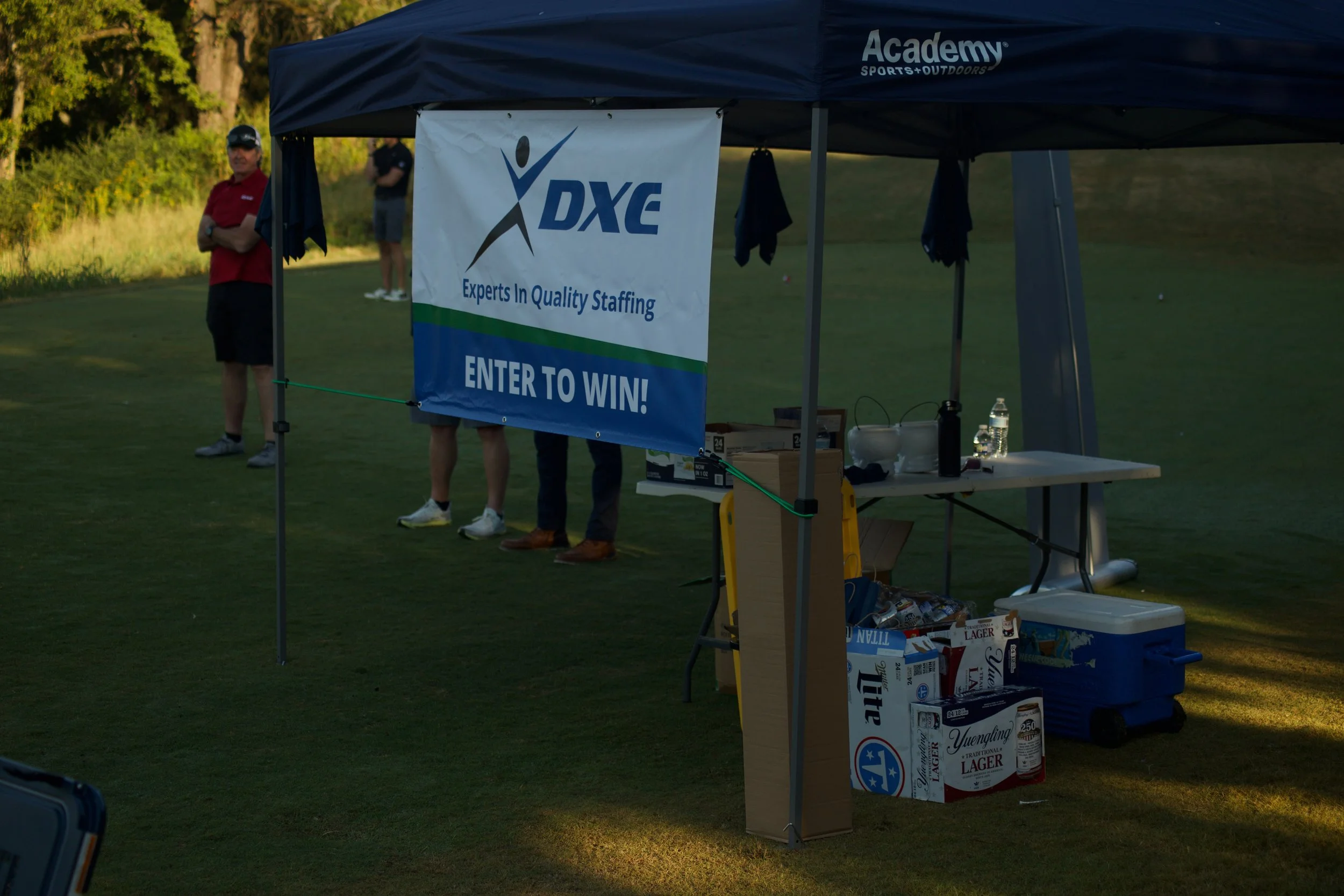An outdoor event tent with a sign for DXE staffing and a sign for entering a contest. People are standing nearby, with trees in the background, on a grassy field at sunset.
