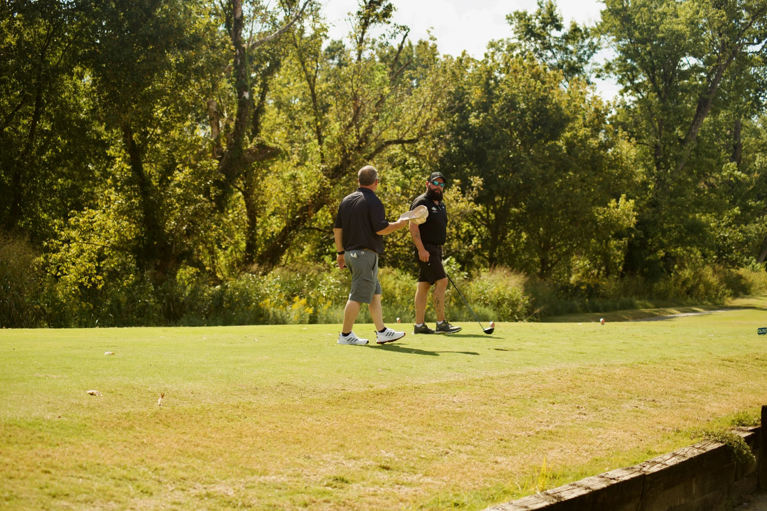 Two men playing golf on a sunny day on a golf course, with trees in the background.