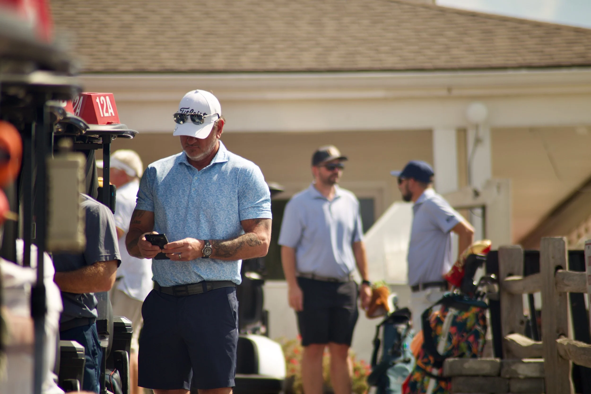 Man wearing sunglasses and a white hat with 'Titleist' logo, looking at his phone at outdoor event with several other people, some wearing hats and sunglasses, and golf equipment.