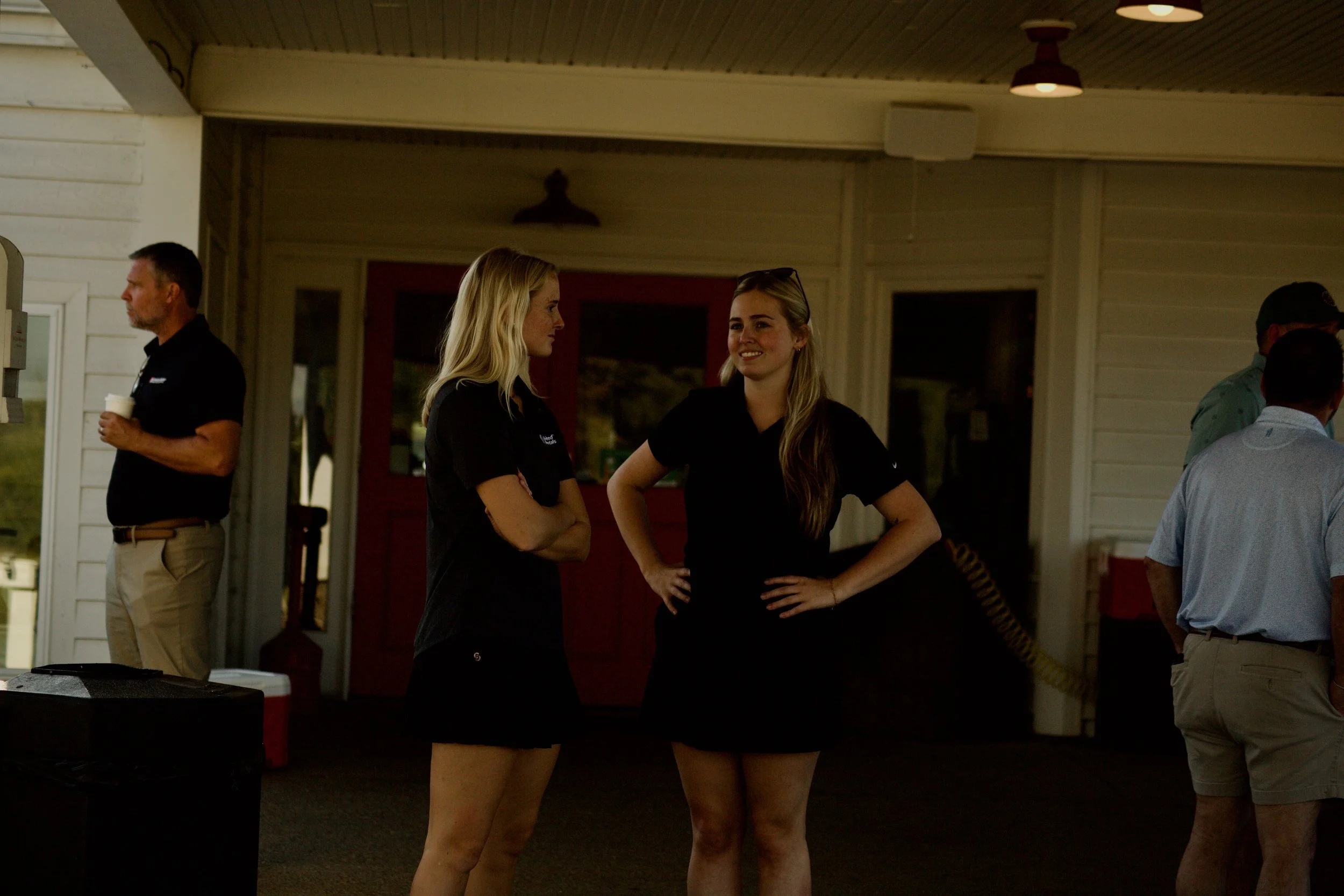 Two women with blonde hair wearing black shirts standing and talking to each other at an outdoor gathering during the evening.