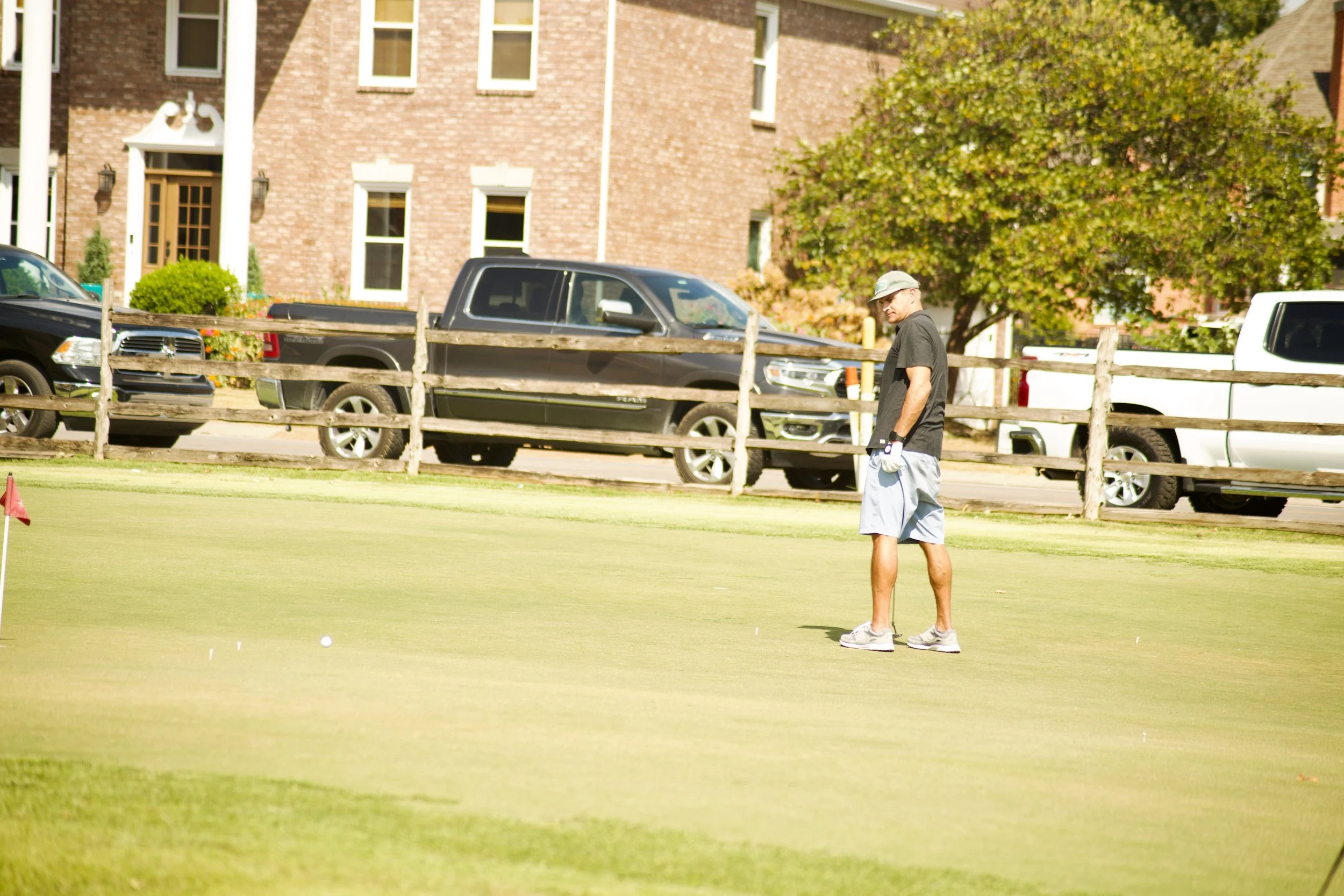 A man standing on a golf course near the hole, looking down at the ground. In the background, there are parked vehicles, a wooden fence, and a brick building with multiple windows and some greenery.