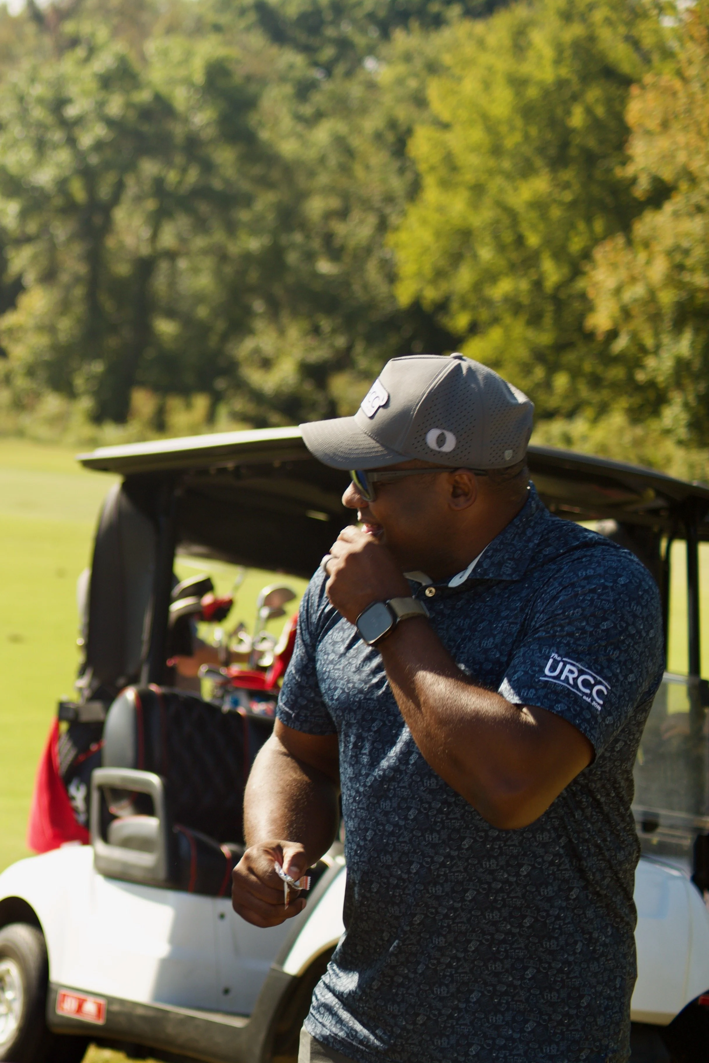 Man wearing a gray baseball cap and a blue patterned polo shirt standing outdoors near a golf cart, with trees in the background.