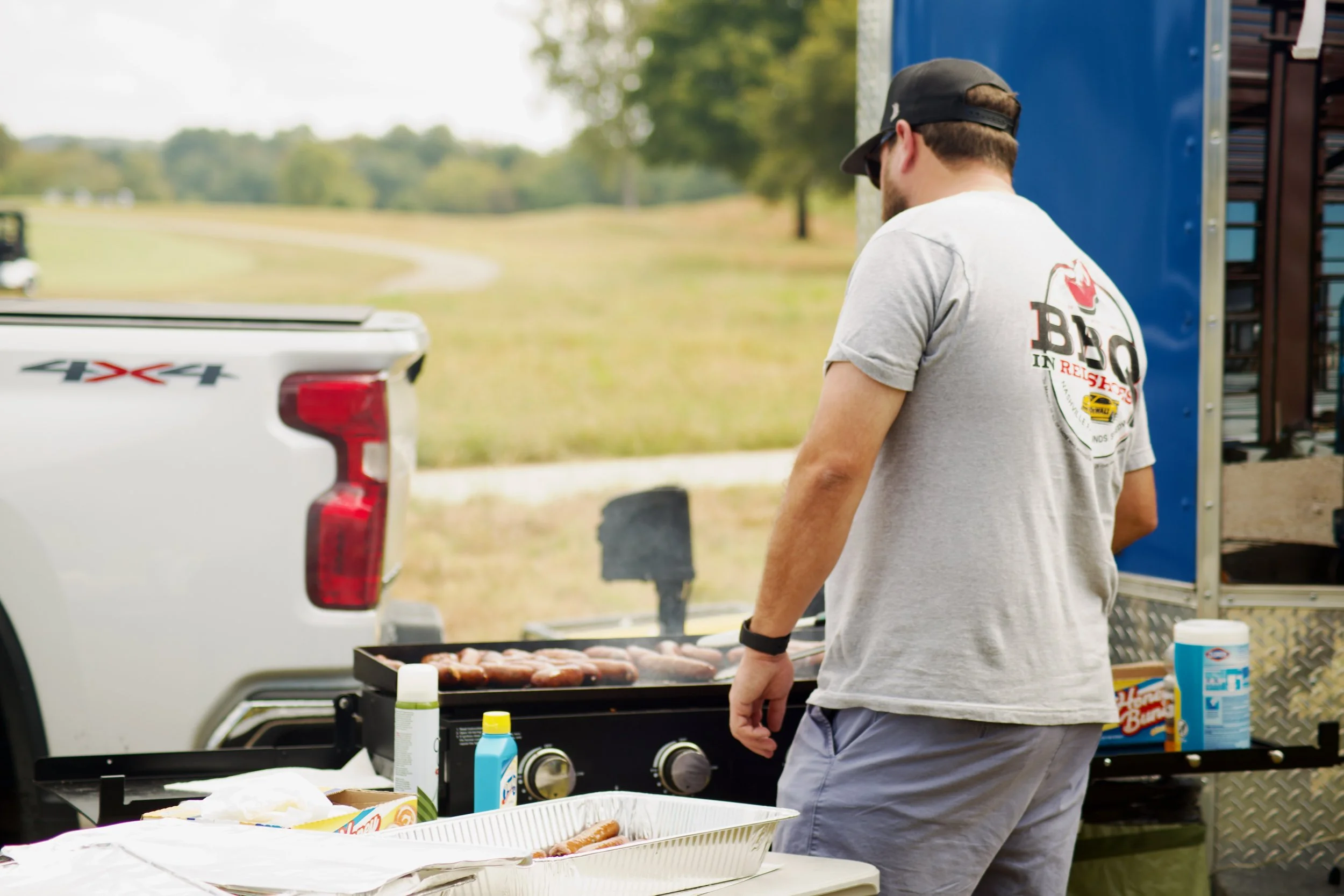 A man wearing a gray t-shirt and a backward black cap is grilling chicken on a barbecue grill outdoors near a pickup truck. The man has a beard and sunglasses. There are condiments and packaging on a table in front of him. It appears to be a casual o
