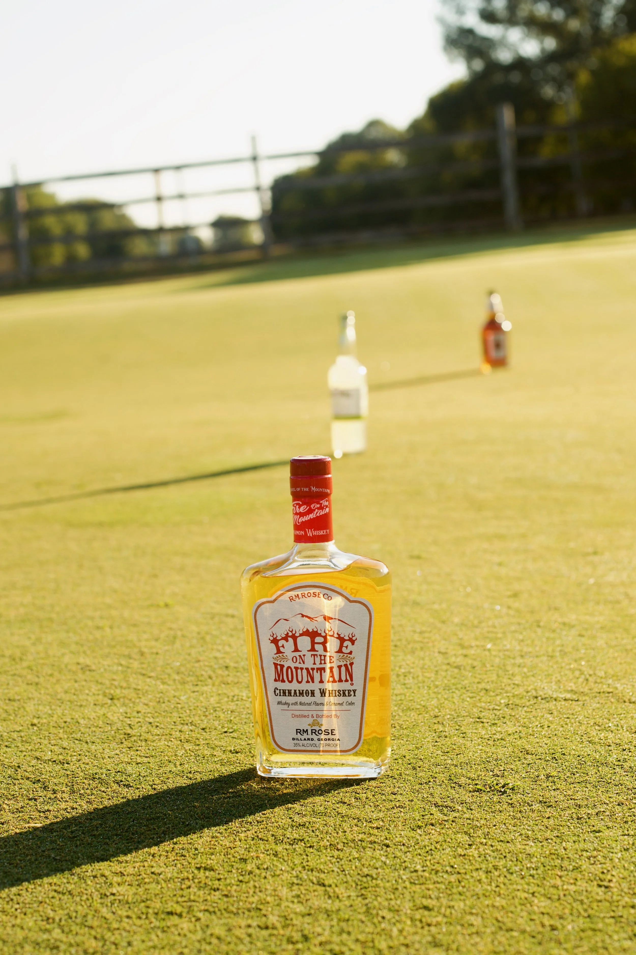 Front view of a bottle of Fire on the Mountain cinnamon whiskey placed on a green lawn, with several other bottles in the background, a wooden fence, and trees under a sunny sky.