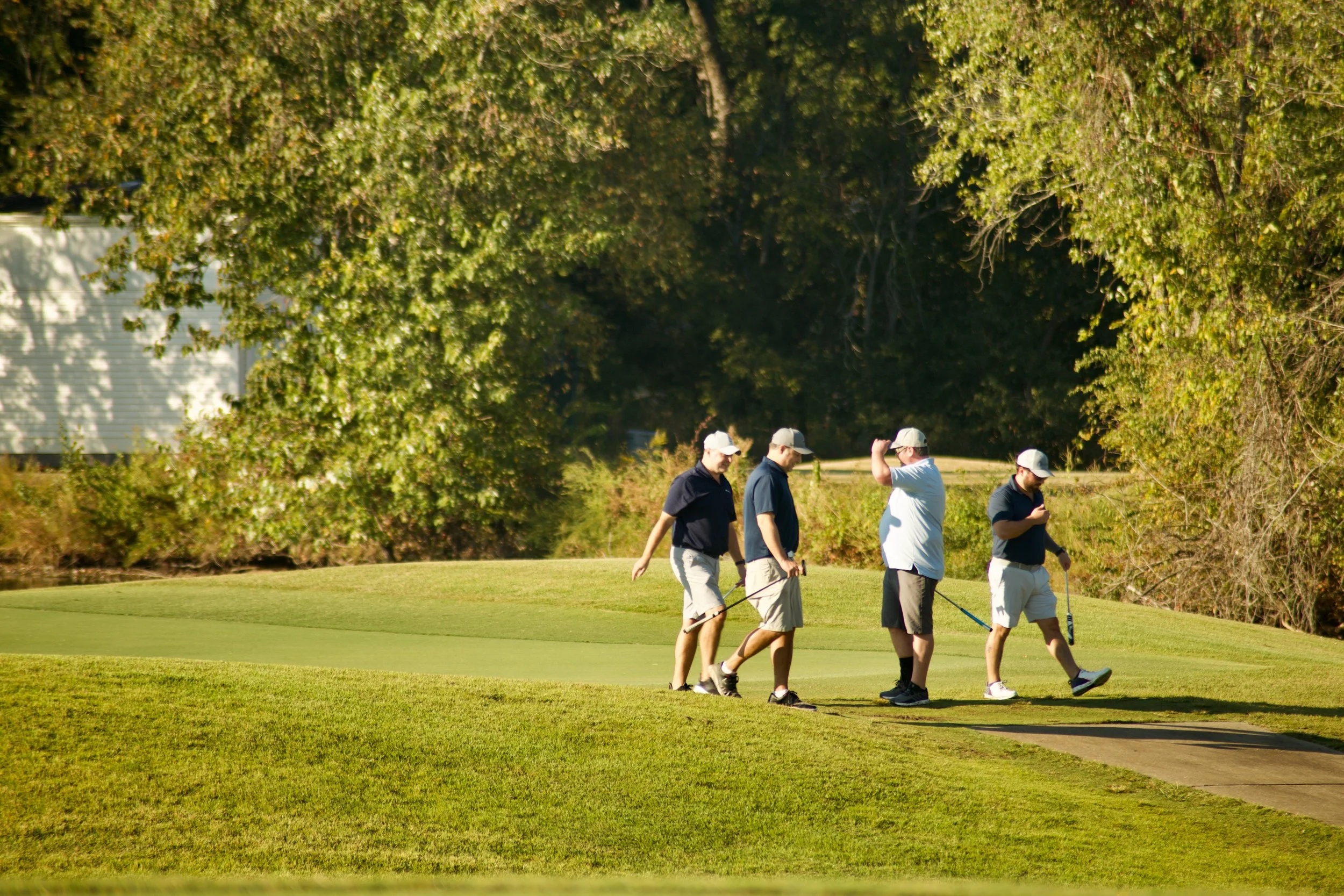 Four people walking on a golf course, talking and using golf clubs, with trees in the background.