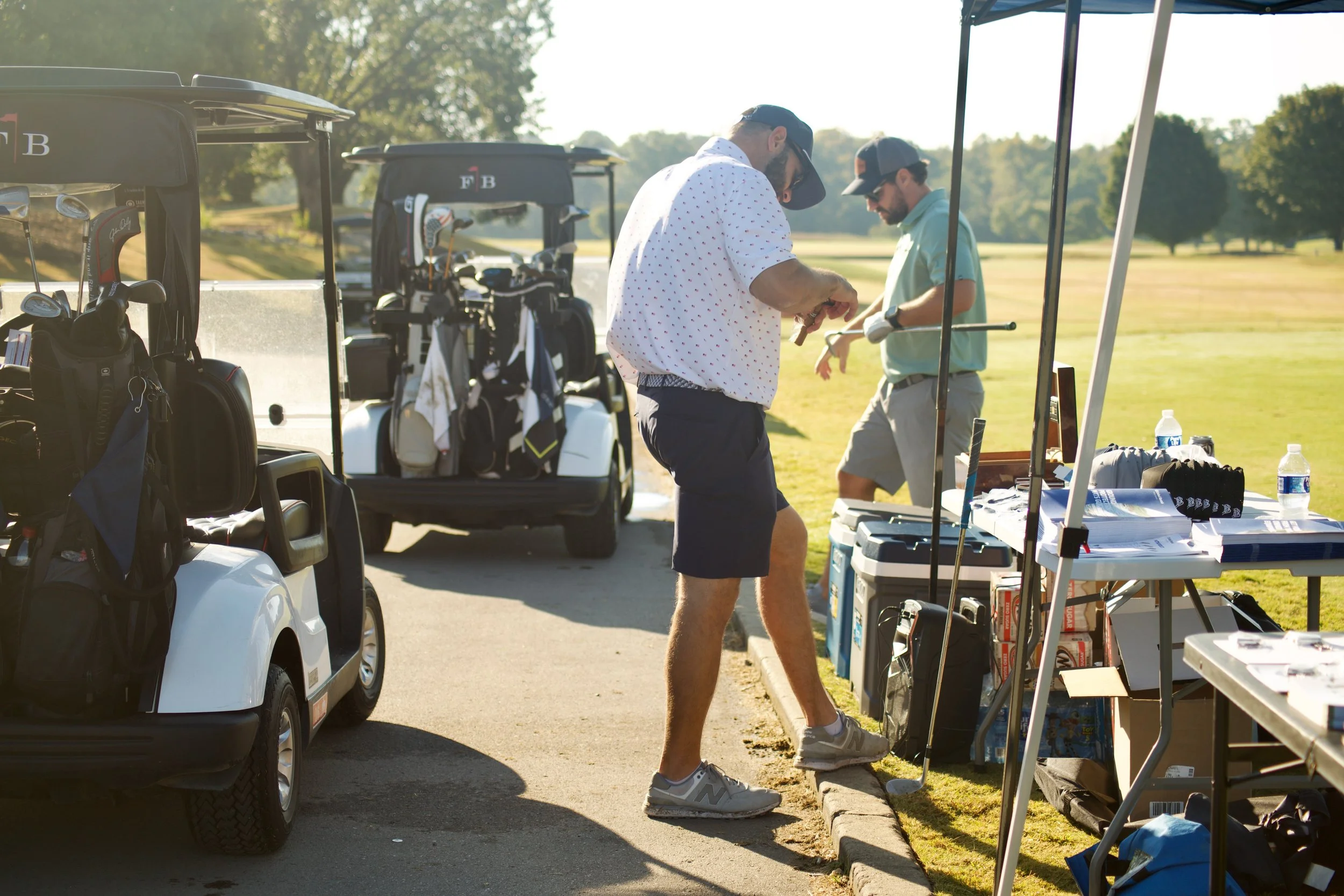Two men at a golf course standing near a table with golf equipment, with golf carts parked nearby, engaged in conversation during sunny weather.