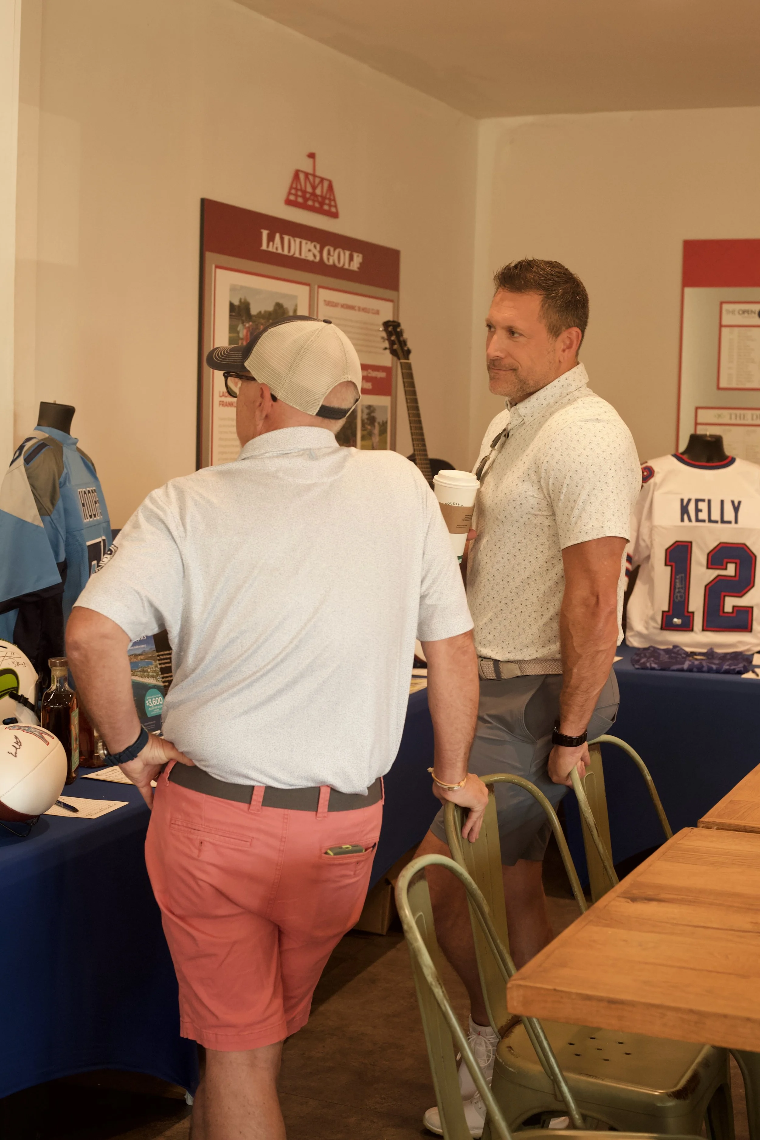 Two men standing at a booth with sports memorabilia, including a Cowboys jersey with the name Kelly and the number 12, and a helmet. One man has his back to the camera, wearing pink shorts and a white shirt, while the other holds a coffee cup, wearin
