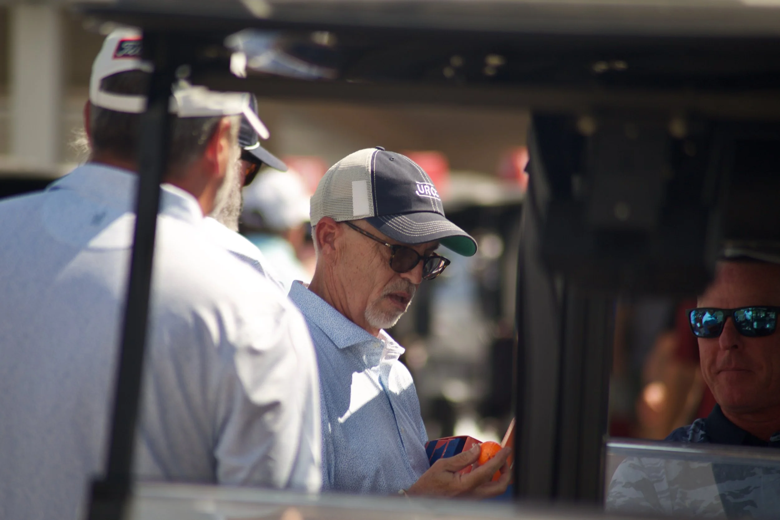 A group of people, mostly older men, standing outdoors, with one person in the center wearing a blue cap and glasses, seemingly looking at a smartphone or device. The scene appears to be in a sunny environment, possibly at an event or gathering.