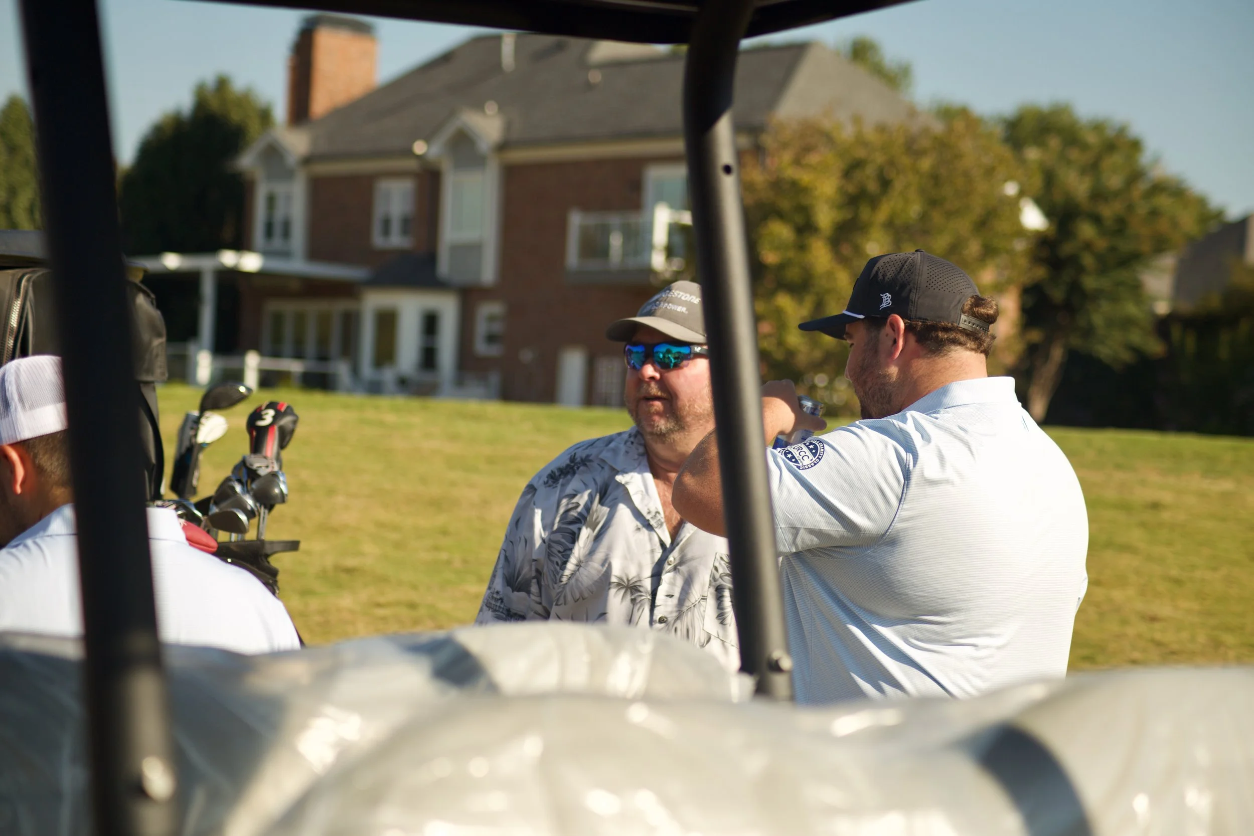 Two men standing beside a golf cart, talking outdoors with a house and trees in the background.
