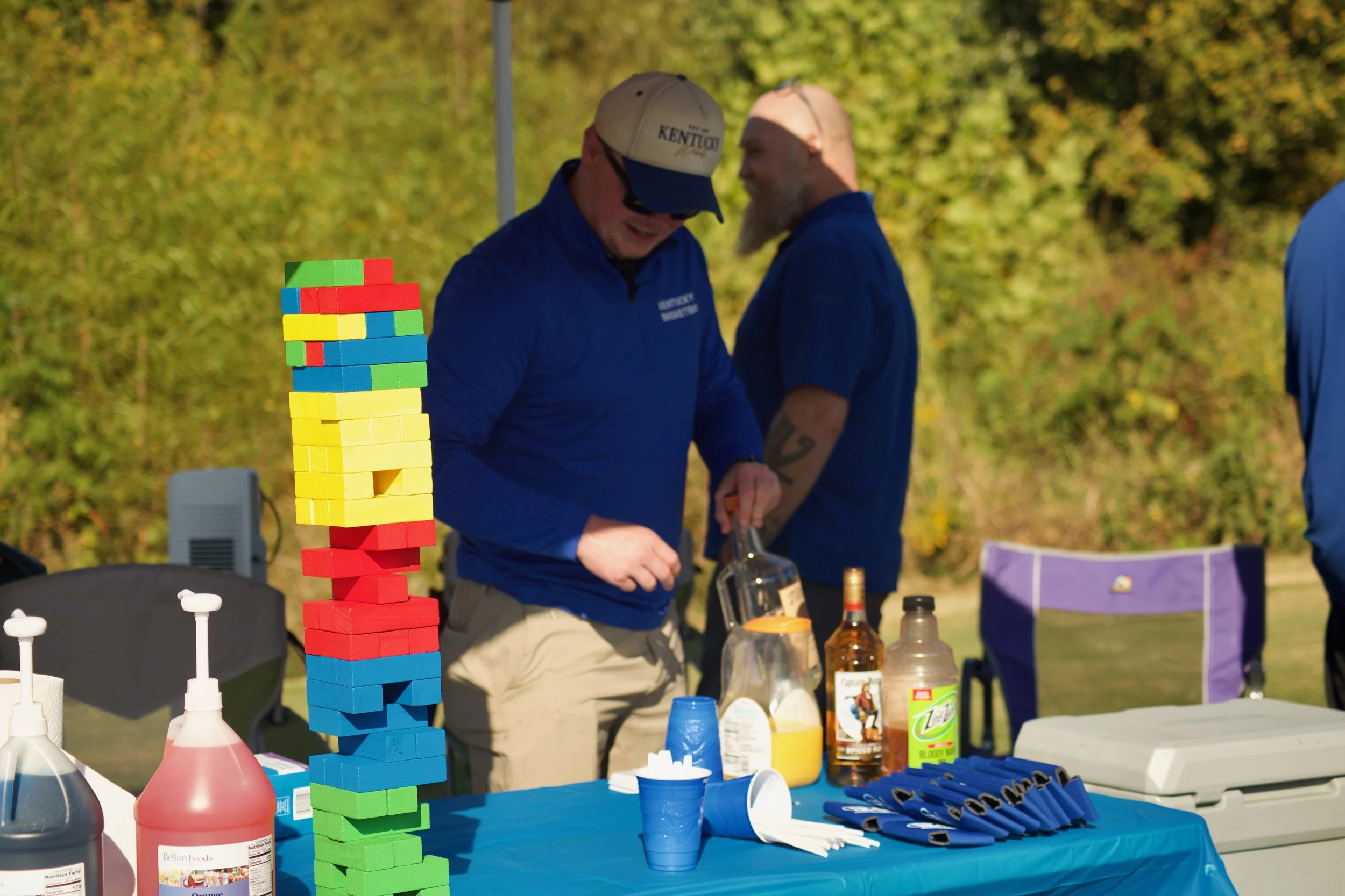 Two men in blue shirts and beige hats playing Jenga outdoors, with a colorful Jenga tower, condiments, and promotional materials on a table.