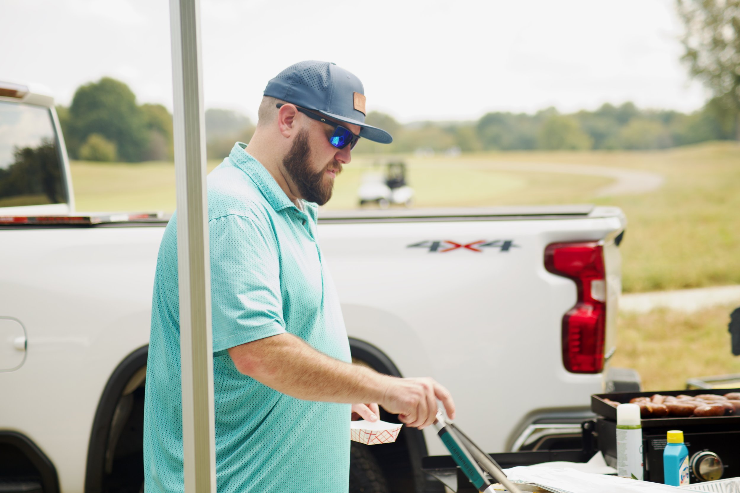 A man with a beard wearing a gray baseball cap, with sunglasses and a light blue shirt, is grilling food outdoors near a white pickup truck with a 4x4 decal, in a grassy area with trees in the background.