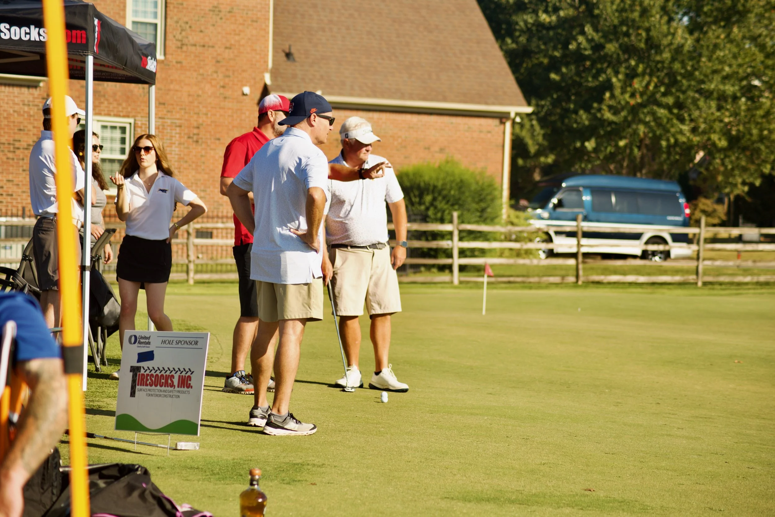Group of people playing golf on a sunny day at a golf course, with a brick building and trees in the background.
