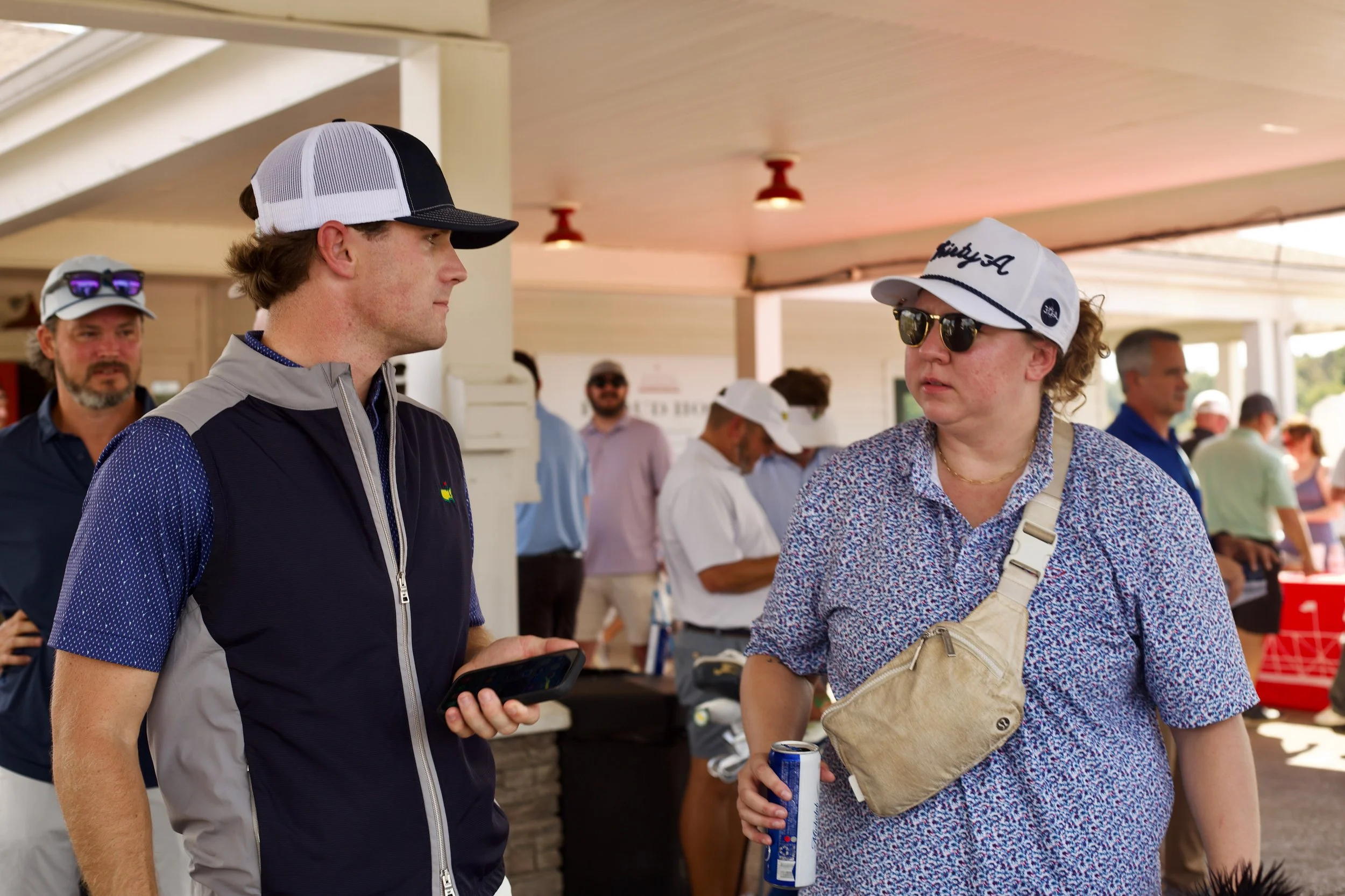 Two people, a man and a woman, are engaged in conversation at an outdoor event. The man wears a white and black trucker hat, a blue and gray vest, and is holding a cellphone. The woman wears glasses, a white cap with text, a patterned shirt, and carr